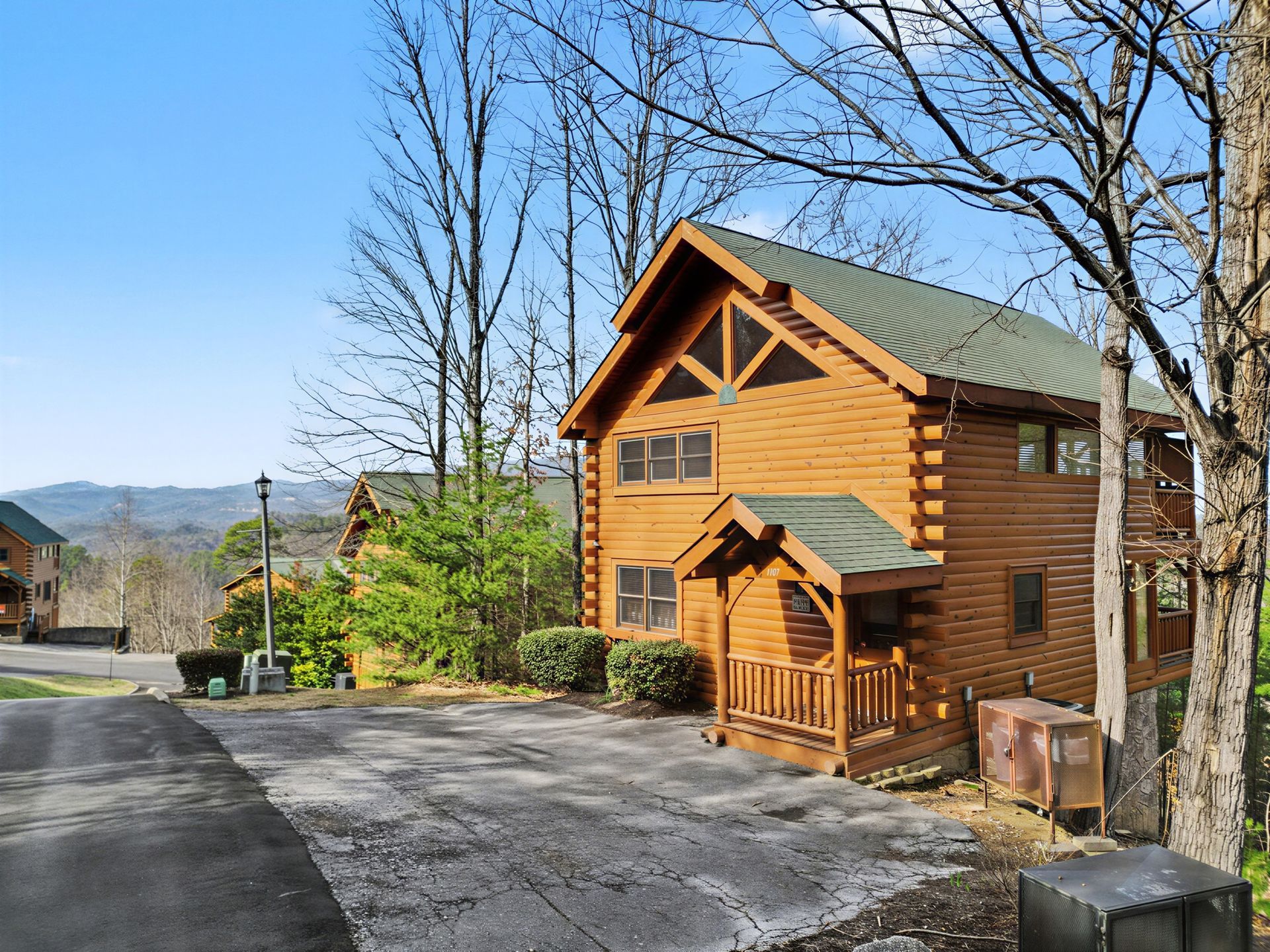 Log cabin with a small porch, sitting in a driveway with trees and mountains in the distance.