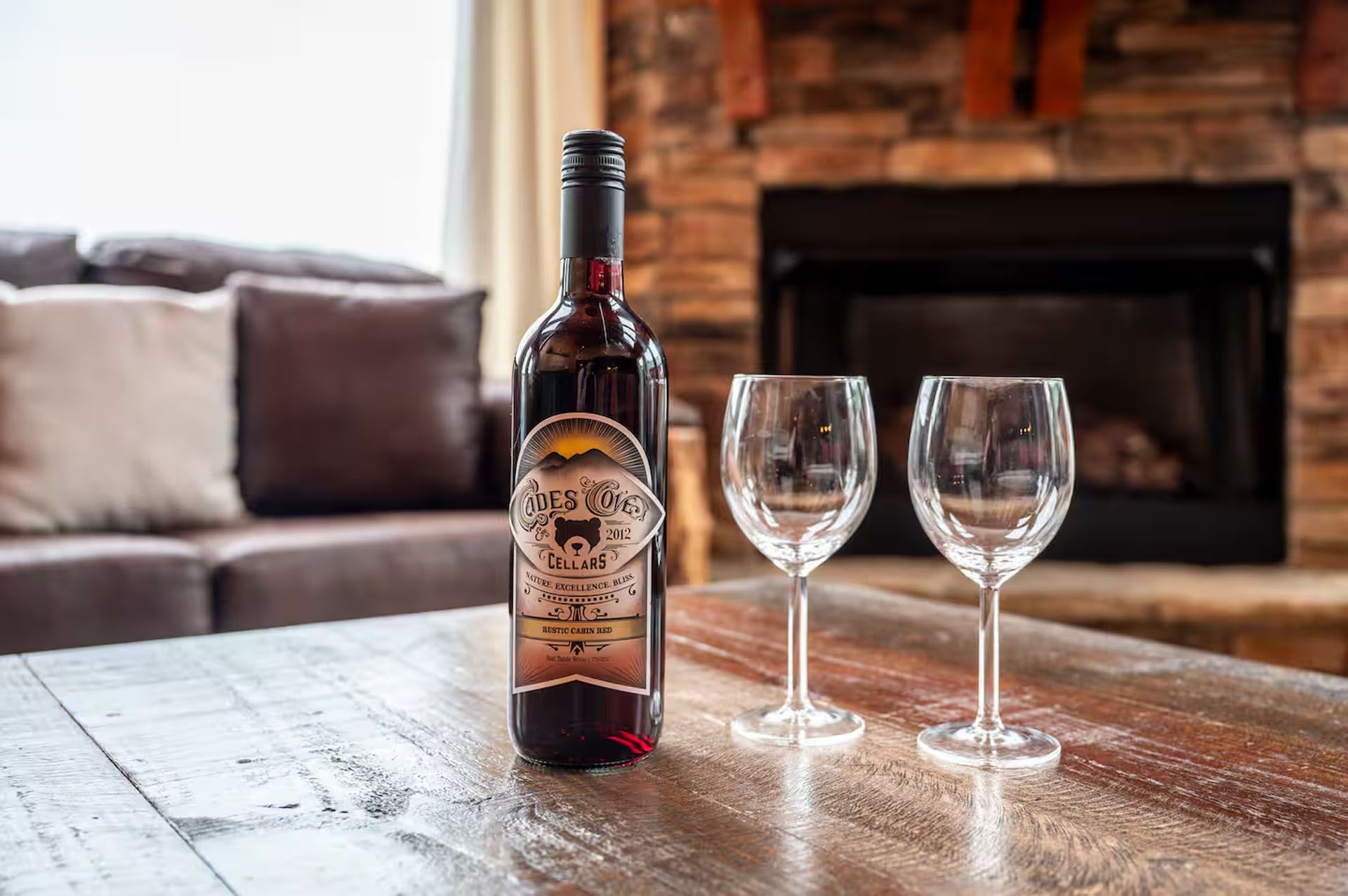 Wine bottle with two glasses on a wooden table in front of a couch and fireplace.