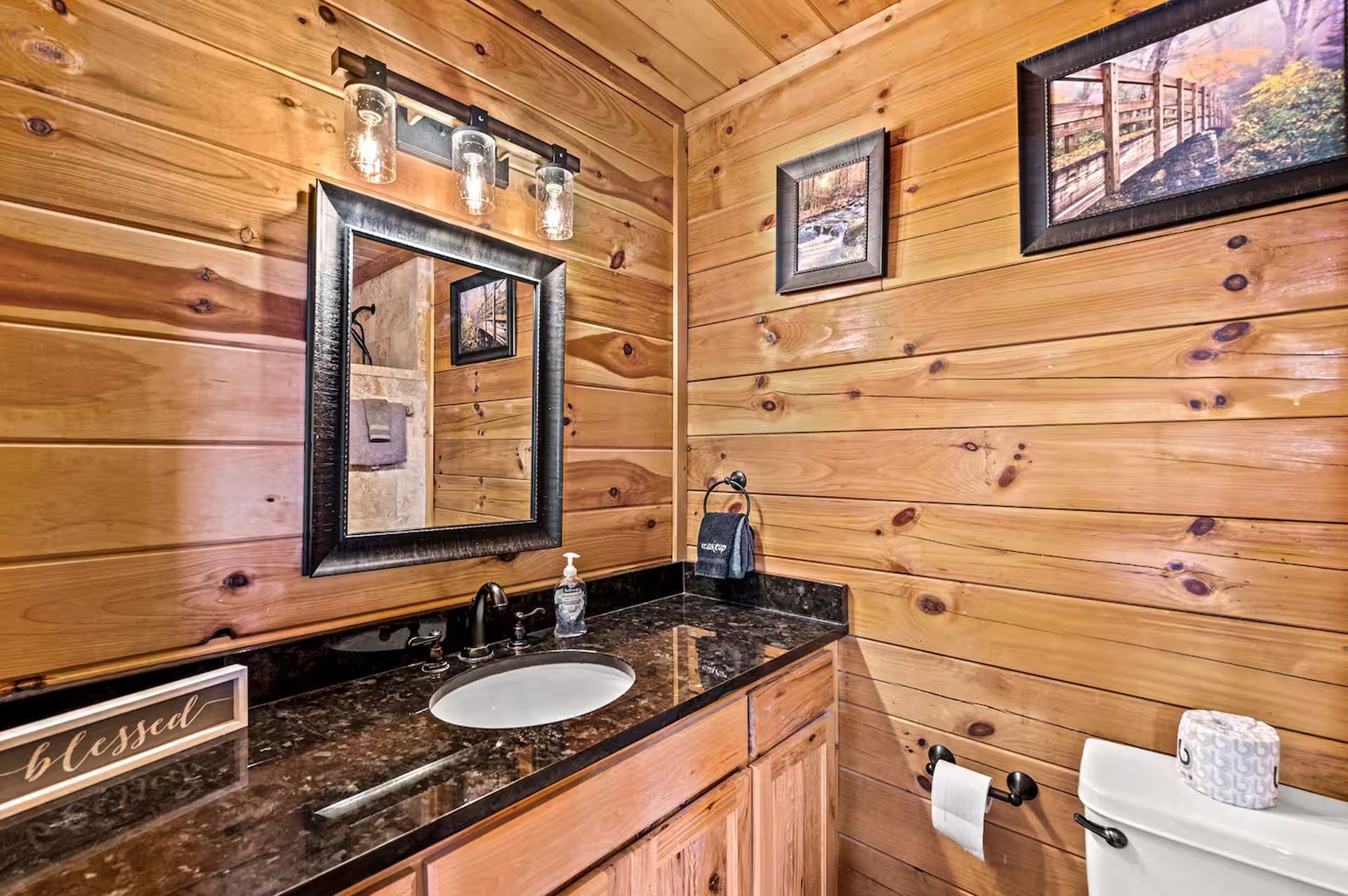 Bathroom with wood-paneled walls, black countertop, mirror, sink, and toilet. Decorative items and framed art are present.