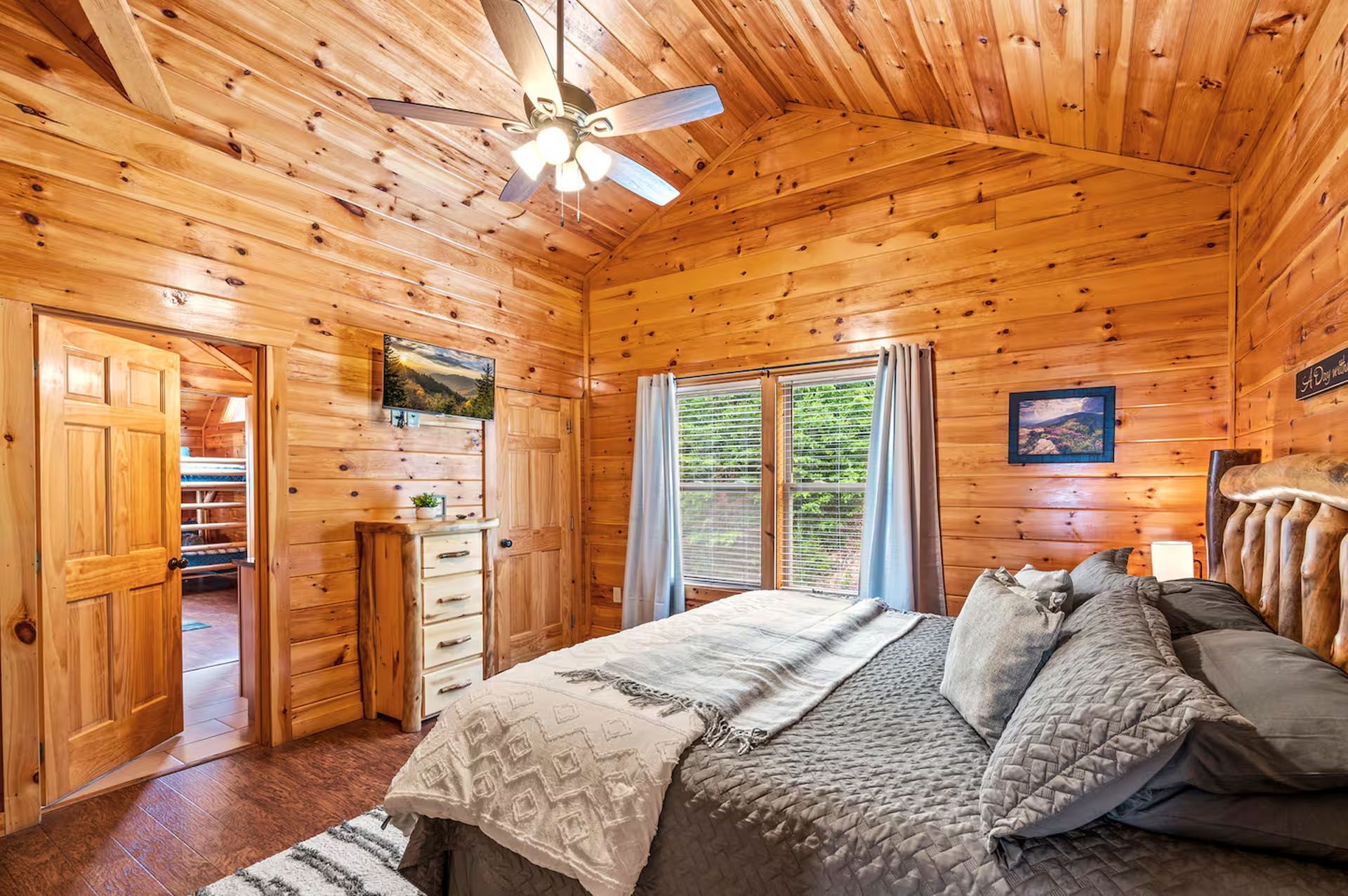 Bedroom with wood paneling, king bed, TV, window with curtains, and a door leading to bunk beds.
