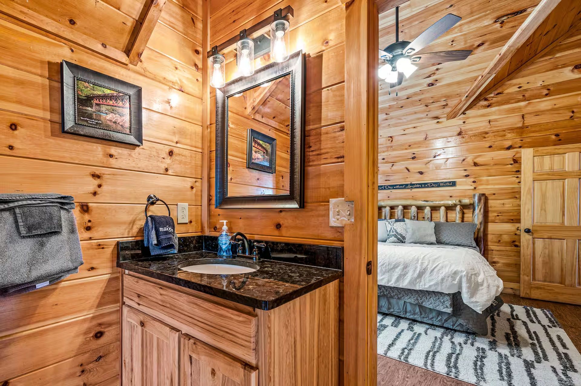 Bathroom and bedroom in a log cabin. Vanity with black countertop, mirror, and door to bedroom with bed.
