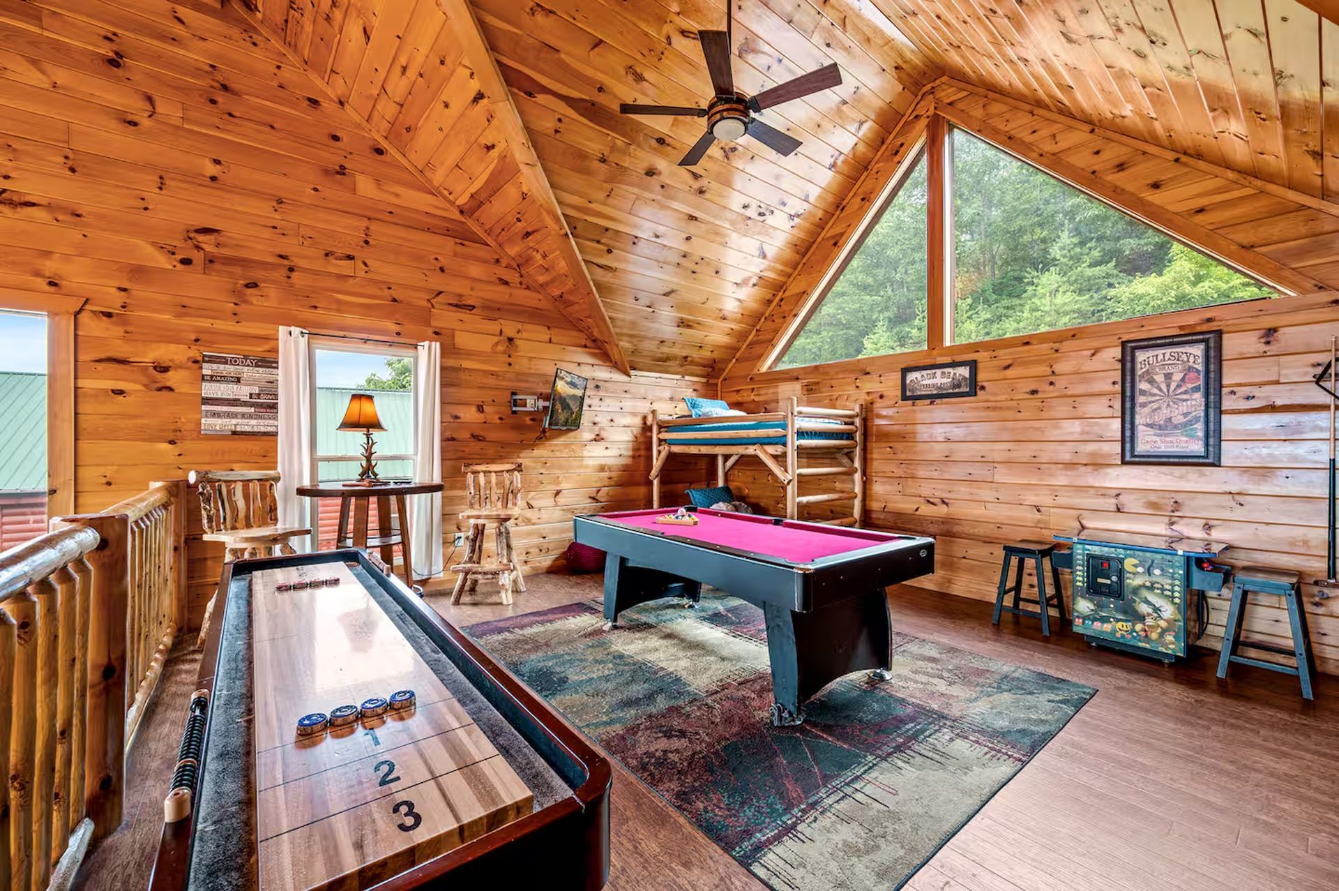 Interior of a wooden-paneled loft with a pool table, shuffleboard, bunk beds, and a large window.