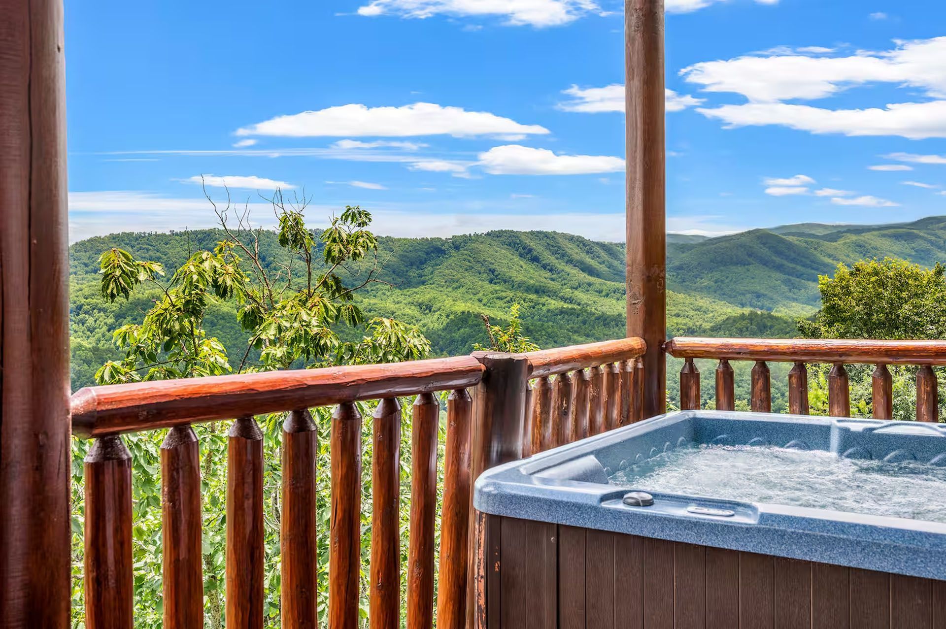 Hot tub on a cabin deck overlooking green mountains under a blue sky with clouds.