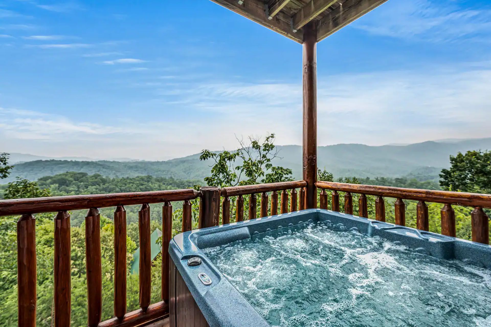 Hot tub on a cabin deck overlooking mountains, clear blue sky.