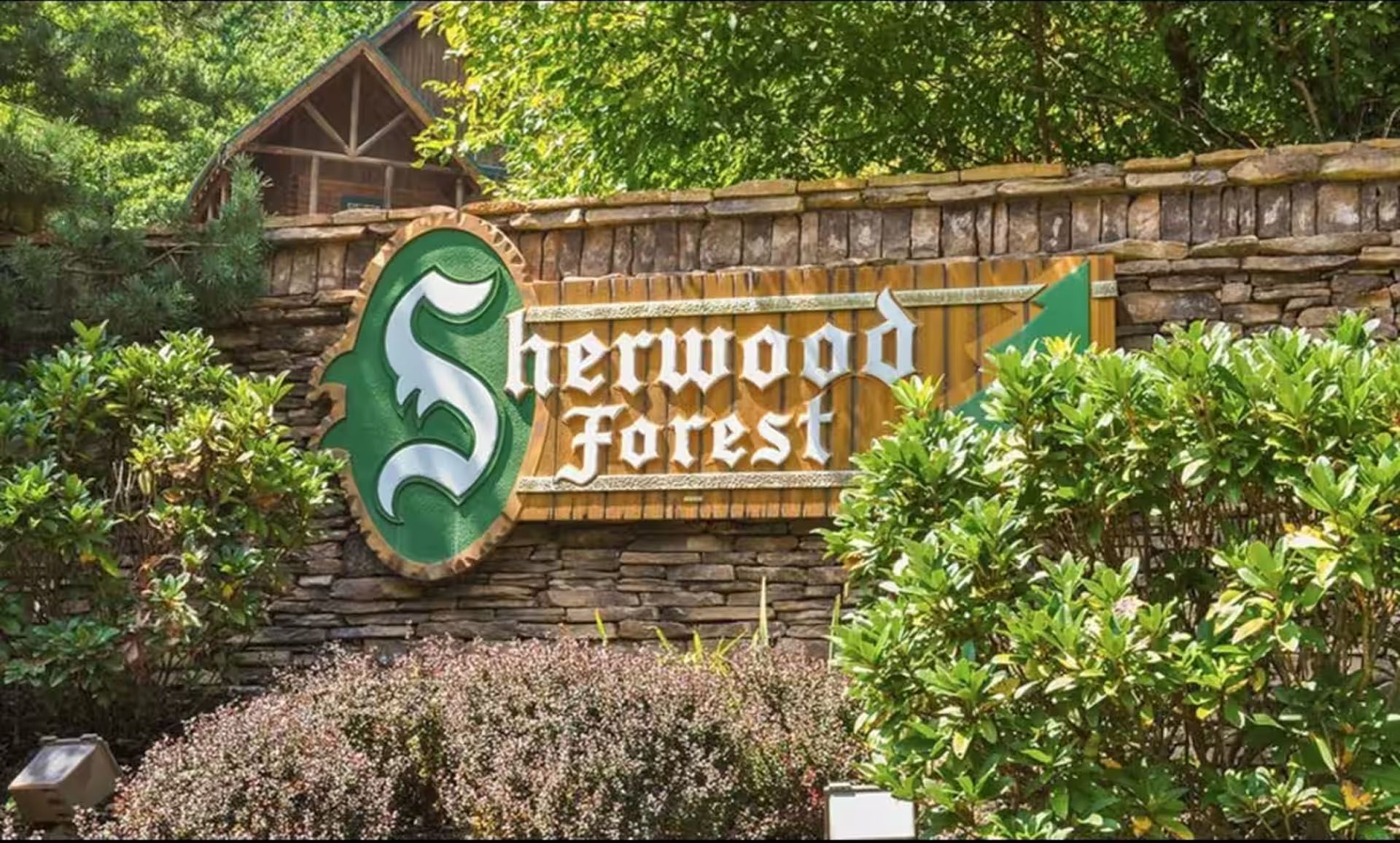 Sign for Sherwood Forest, with green and white emblem on a stone wall, flanked by bushes and trees.