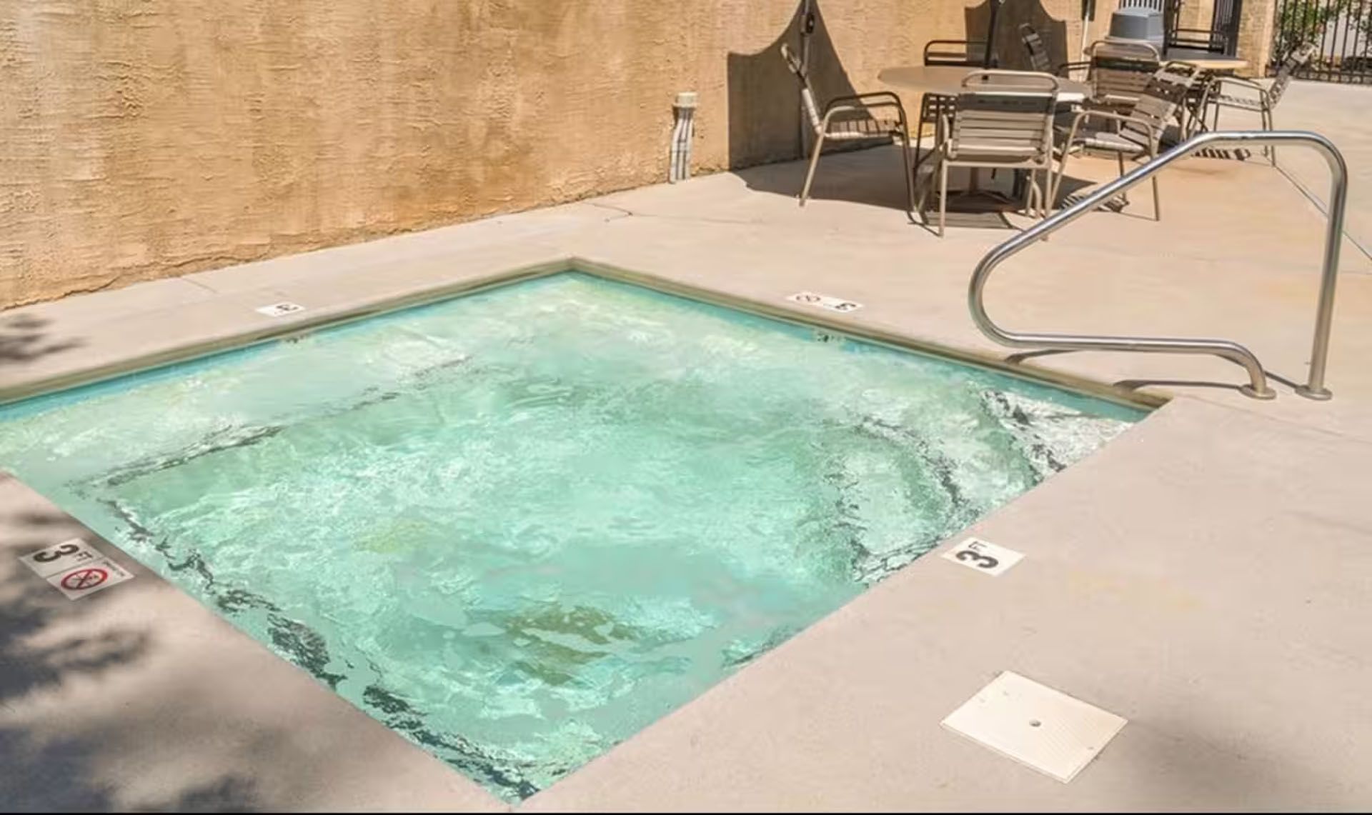 Small, square outdoor hot tub with light blue water, concrete surround, and metal handrail.