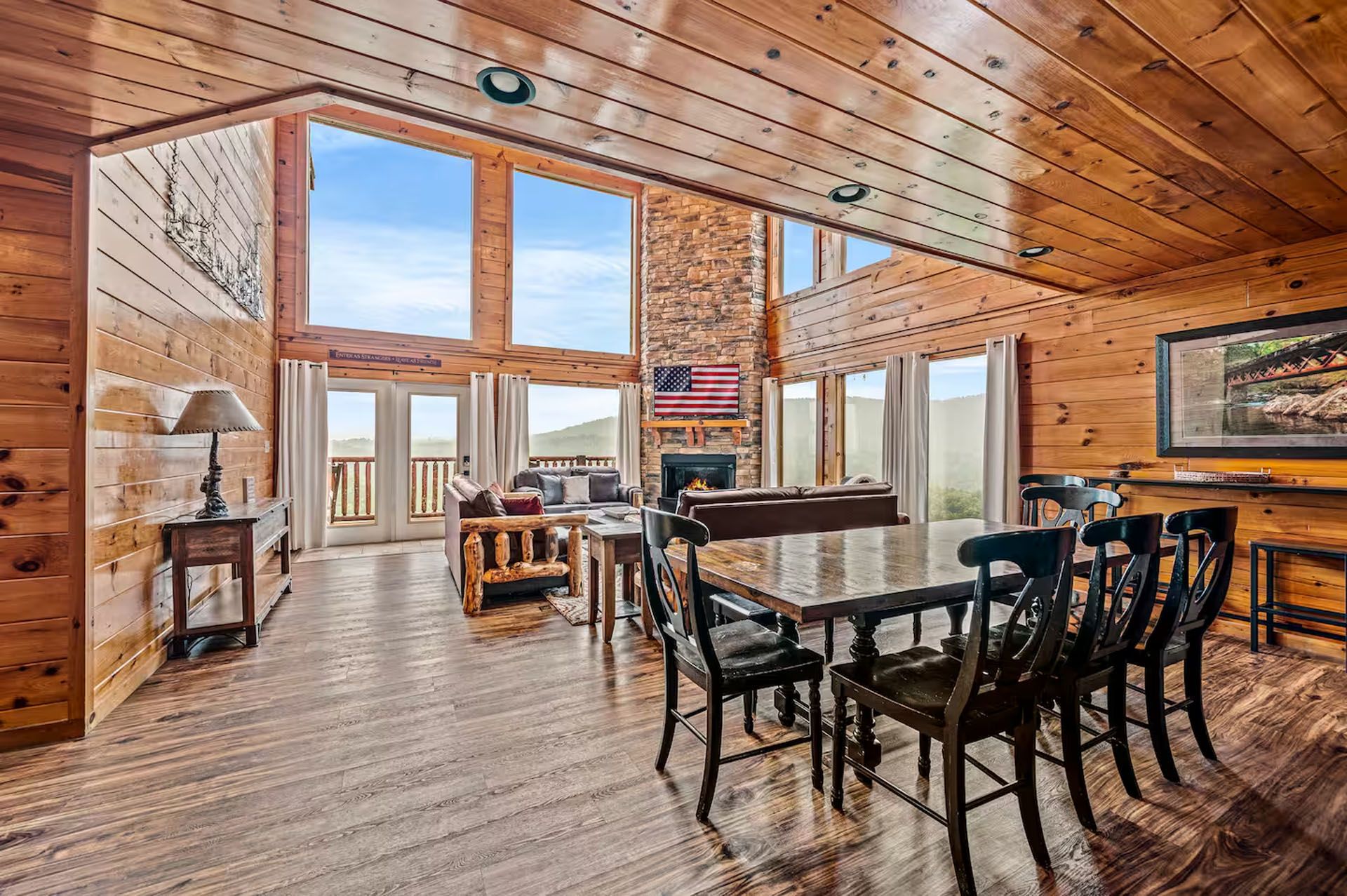 Spacious cabin interior with wooden walls, dining table, and large windows overlooking a landscape.
