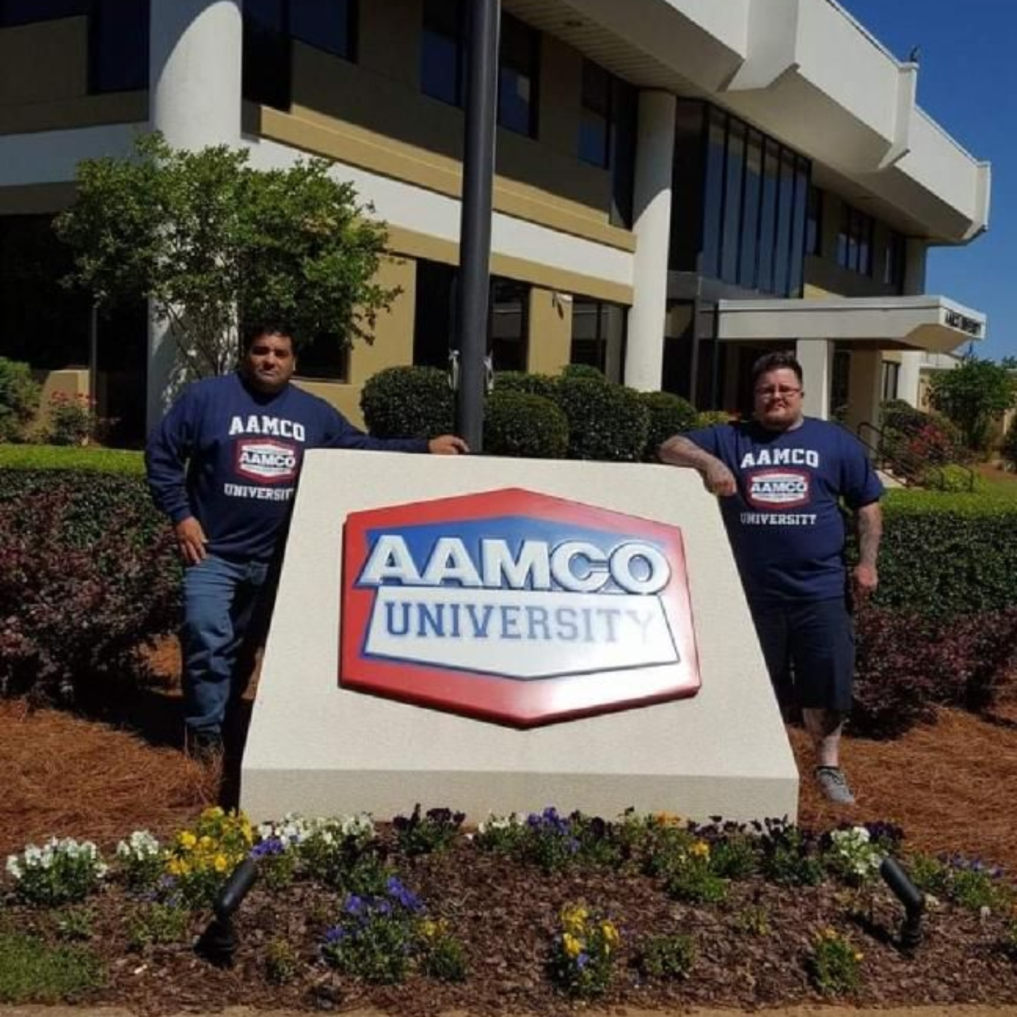 Two people stand beside an AAMCO University sign in front of a building.