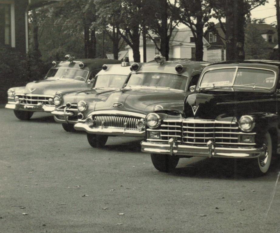 Four vintage ambulances parked in a row, with a black car in front, trees in the background.