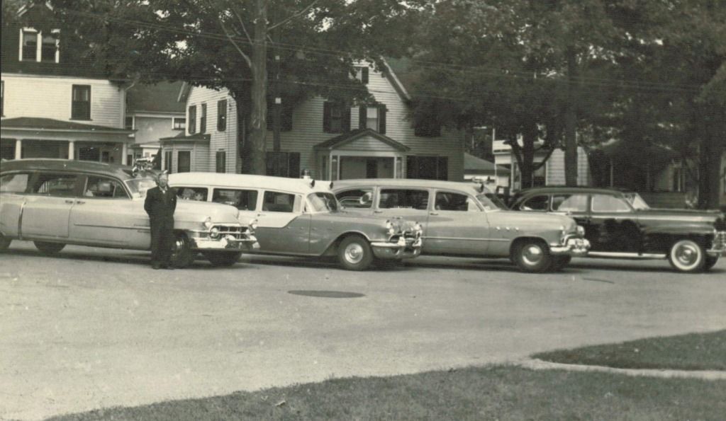 Several vintage cars parked on a street in front of houses; a person stands next to a car.