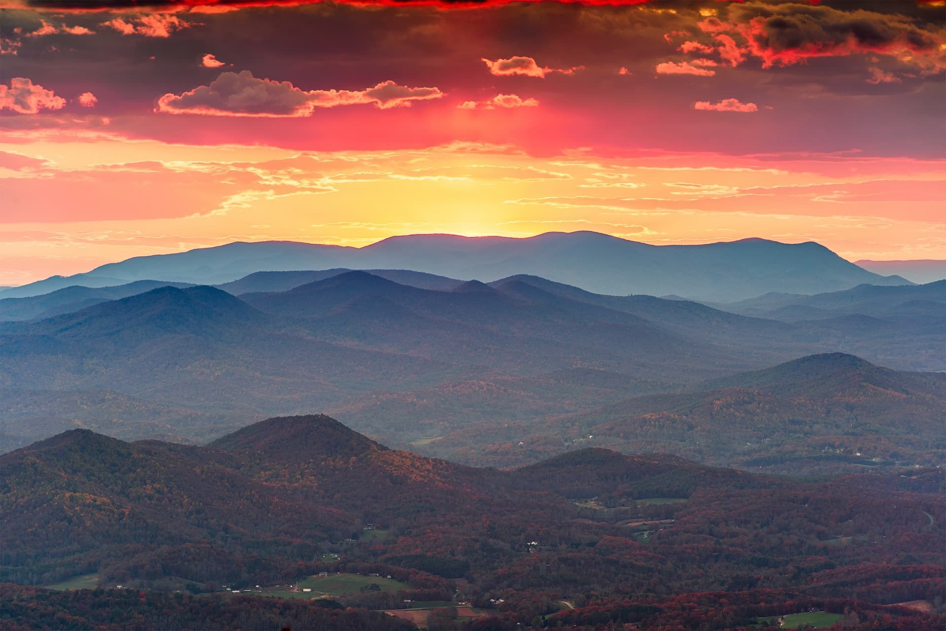 Sunset over a range of blue-hued mountains with hints of trees. Fiery orange and red sky.