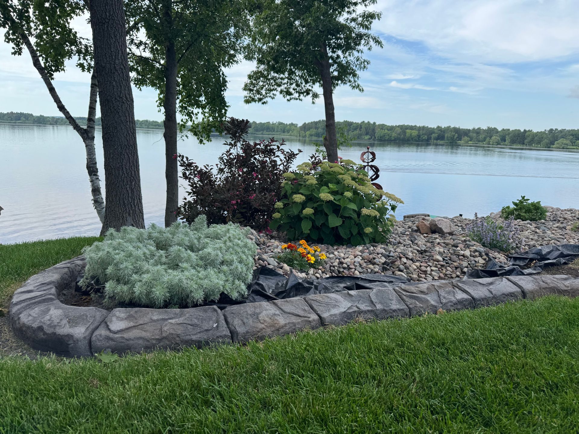 Landscaped lakeside garden with shrubs, flowers, stone edging, and trees overlooking calm water