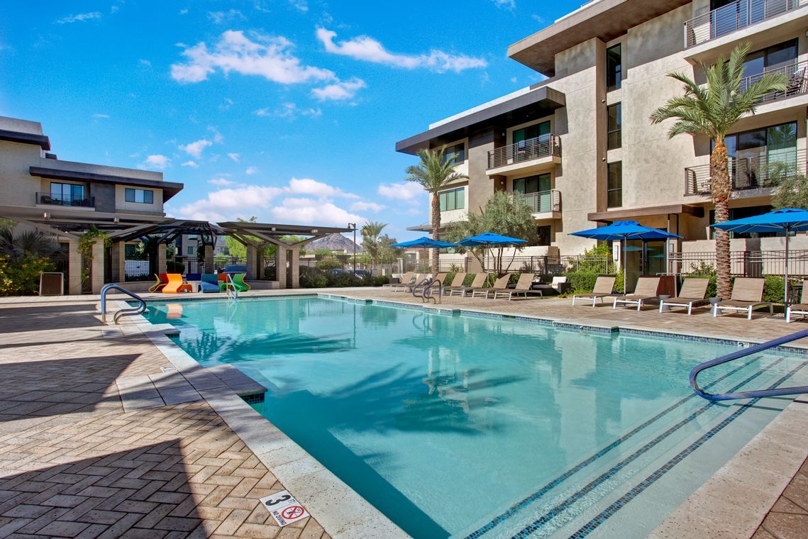 A large swimming pool surrounded by chairs and umbrellas in front of a building.