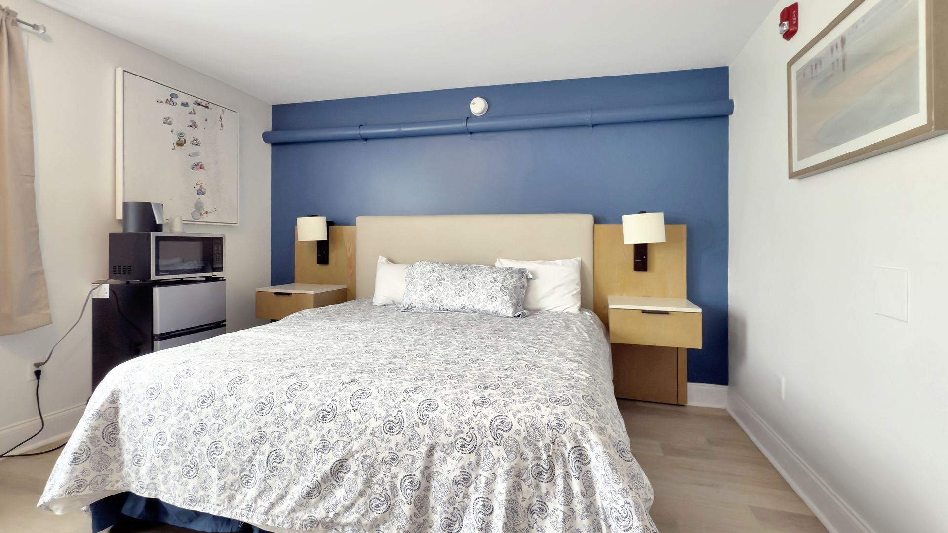 Bedroom with bed, nightstands, blue accent wall, TV, mini-fridge, and artwork.
