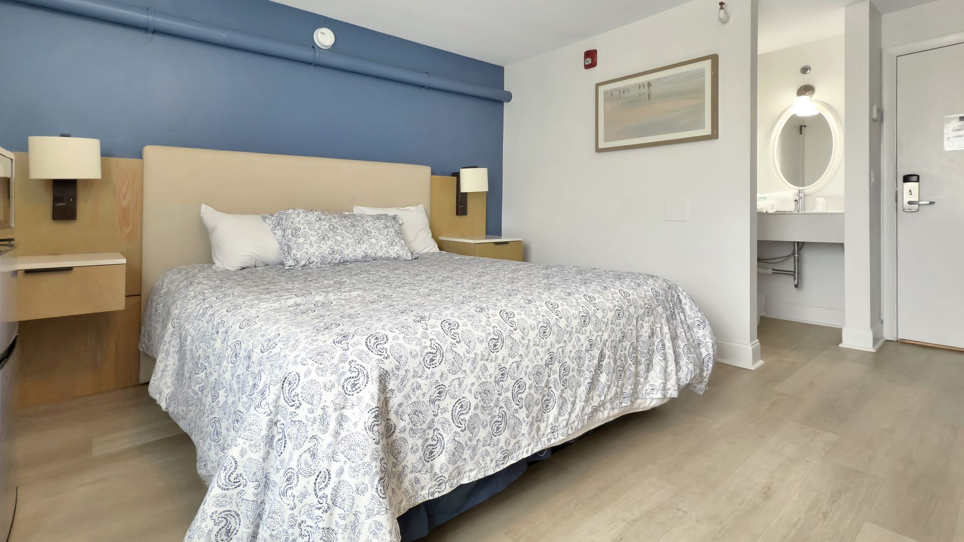 Bedroom with a bed, nightstands, and bathroom visible in the background; blue and neutral color scheme.