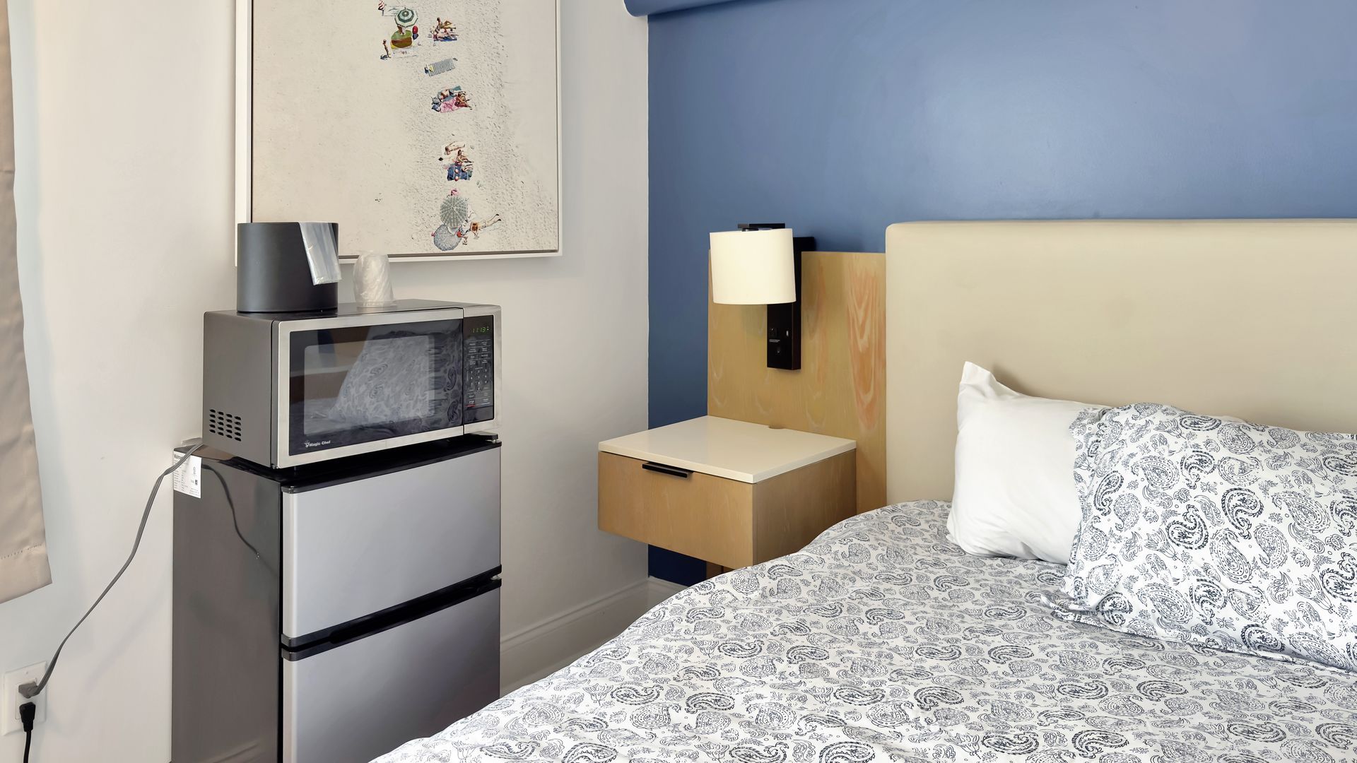 Bedroom with bed, nightstand, microwave, and mini-fridge. Blue accent wall, artwork, and light-colored bedding.