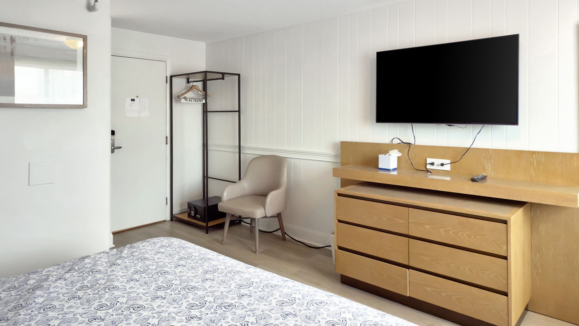 Bedroom with bed, TV, dresser, chair, door, and rack. Neutral colors dominate the space.