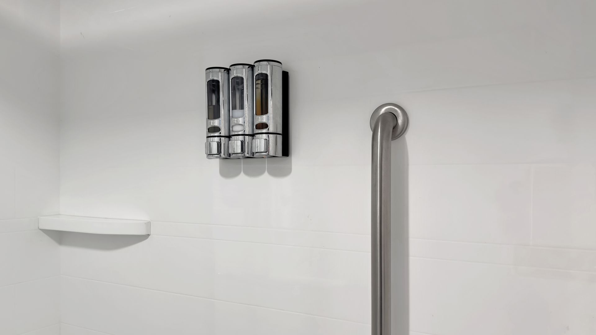 White shower wall with a silver grab bar, soap dispenser, and a soap dish.