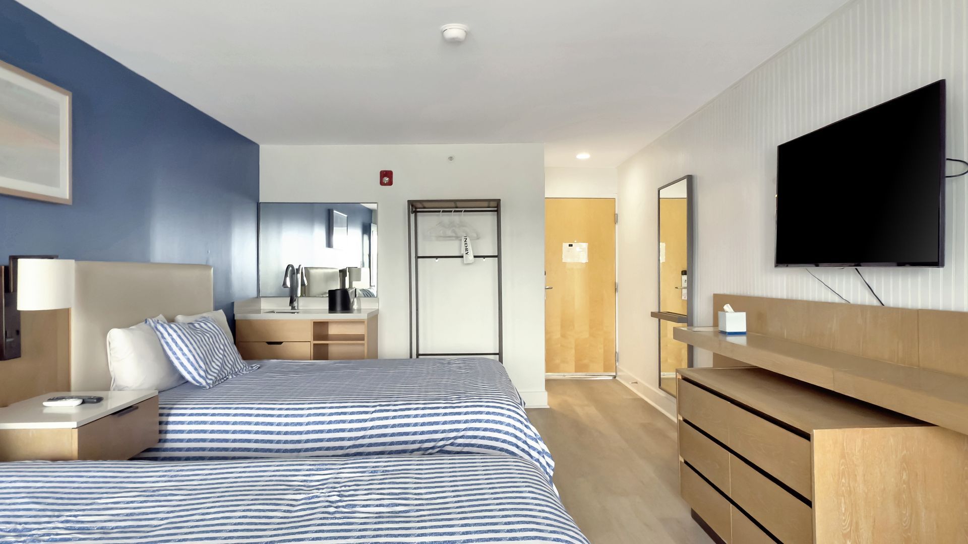Hotel room with two beds, blue and white striped bedding, TV, and light wood furniture.