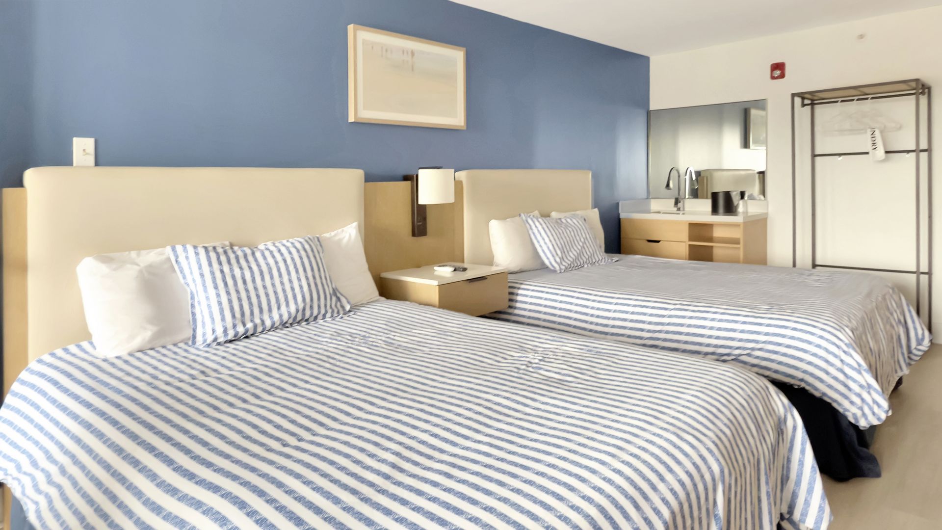 Two beds in a hotel room with blue and white striped bedding, blue wall, and a clothes rack.