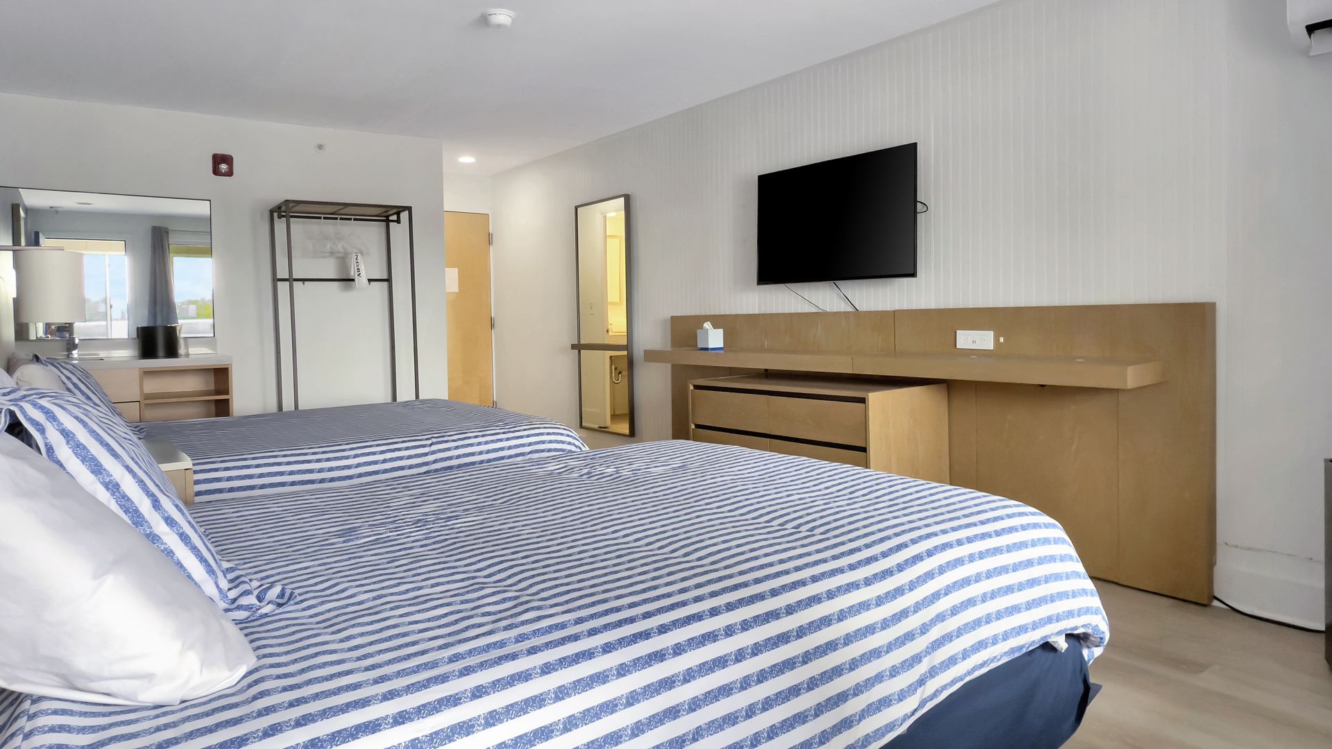 Hotel room with two beds, TV on wall, light wood desk, and striped blue blankets.