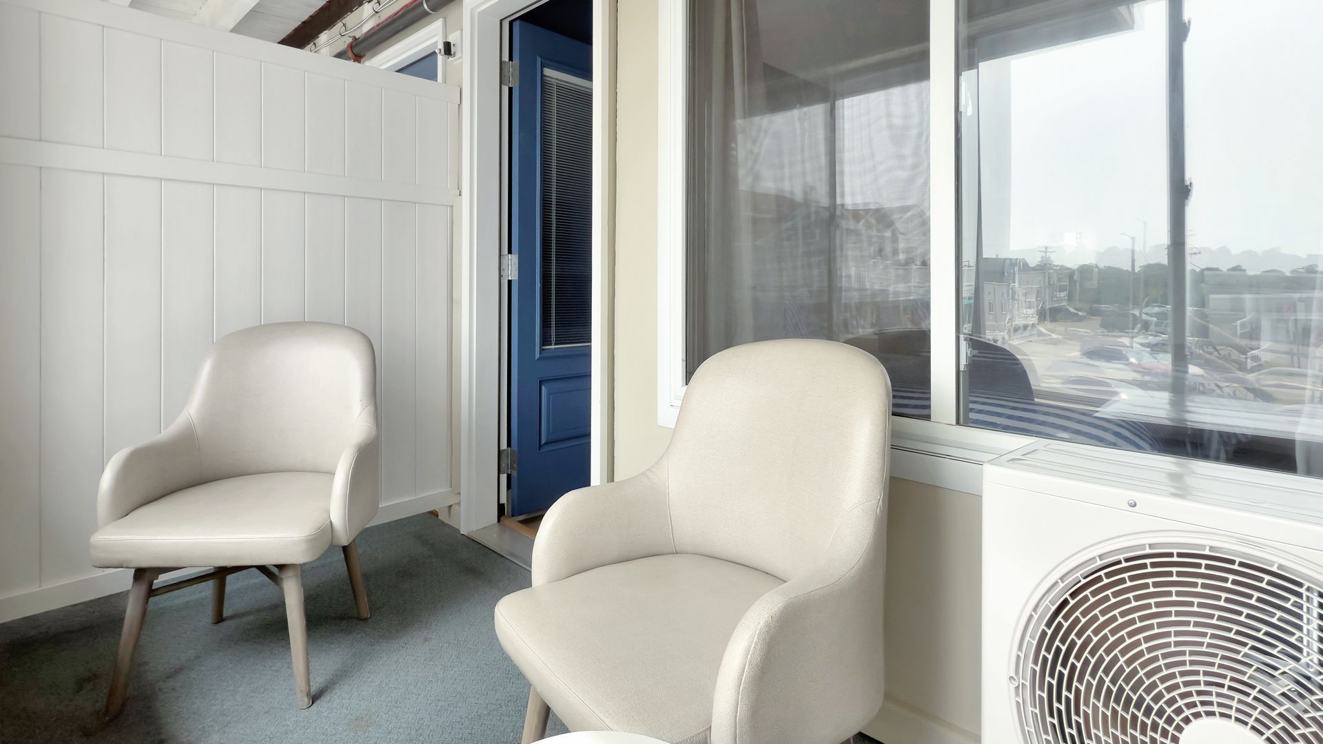 Two cream-colored chairs in front of a window overlooking a cityscape. A blue door is visible behind the chairs.