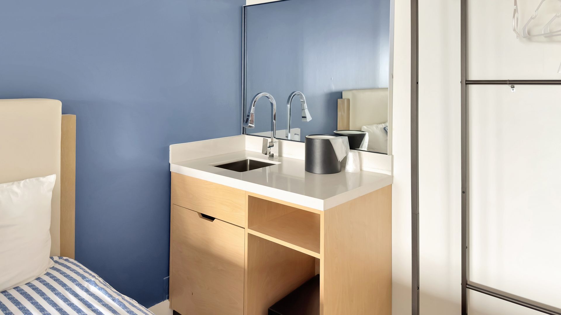Light wood vanity with sink, two-handle faucet, and blue wall. Small mirror on the side and partially visible bed.