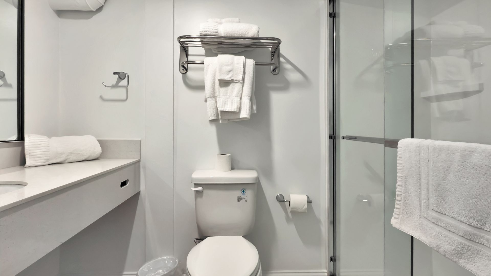 Bright white bathroom with toilet, sink, and shower. Towels hang from a rack.