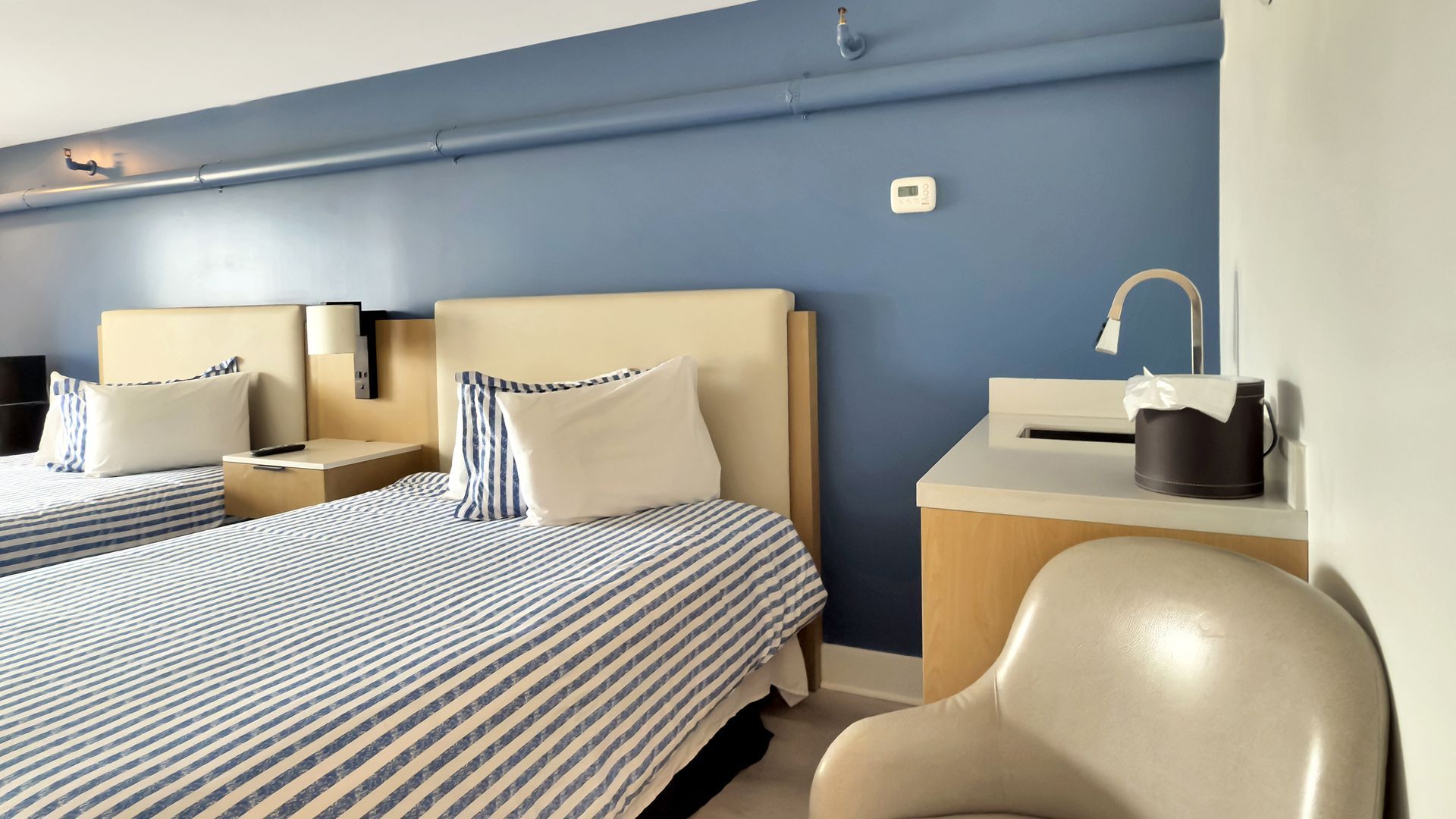 Two beds in a hotel room, blue striped bedding, blue wall, white sink, beige chair.