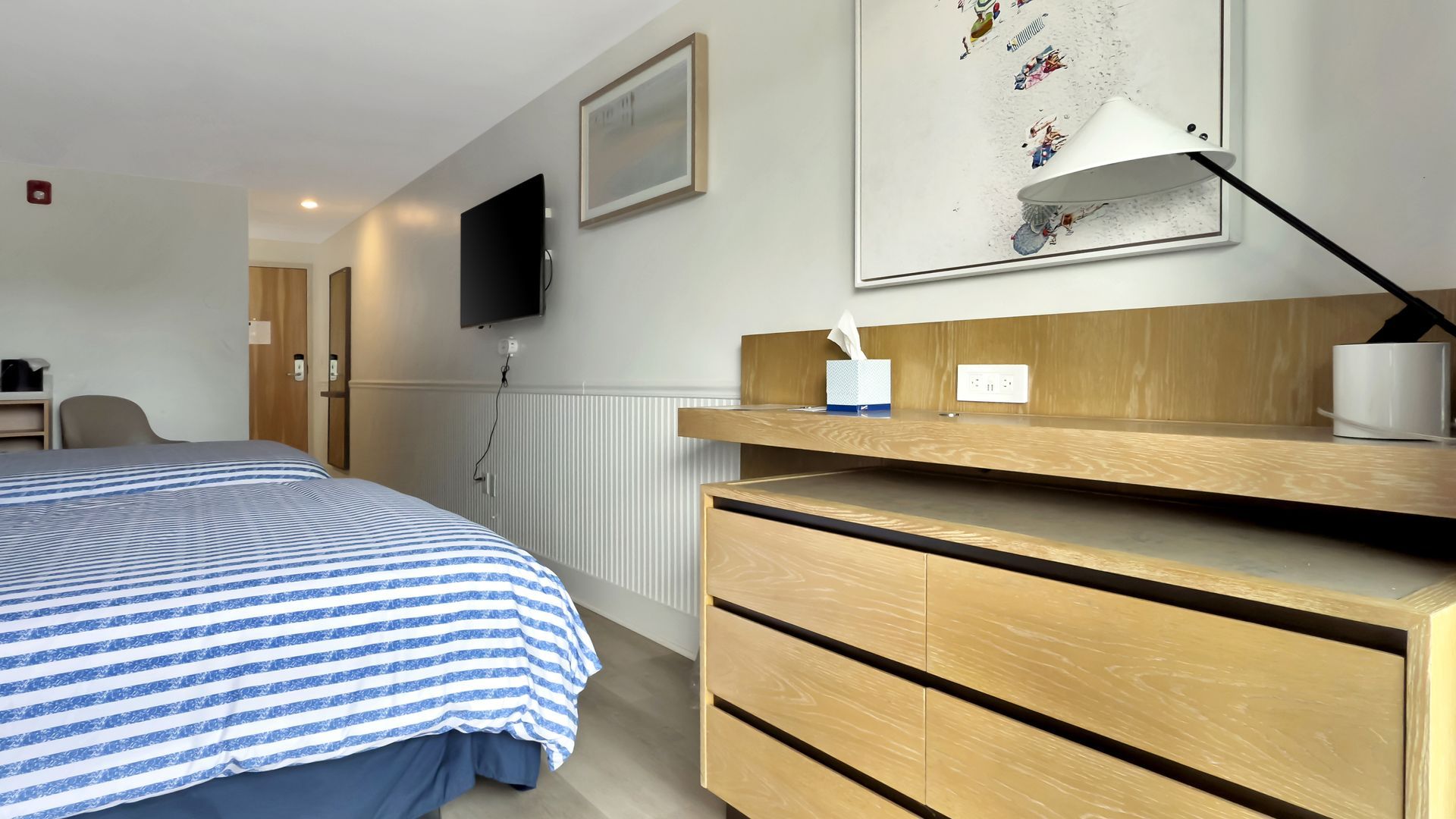 Hotel room with bed, desk, TV, and artwork; white walls, light wood furniture.