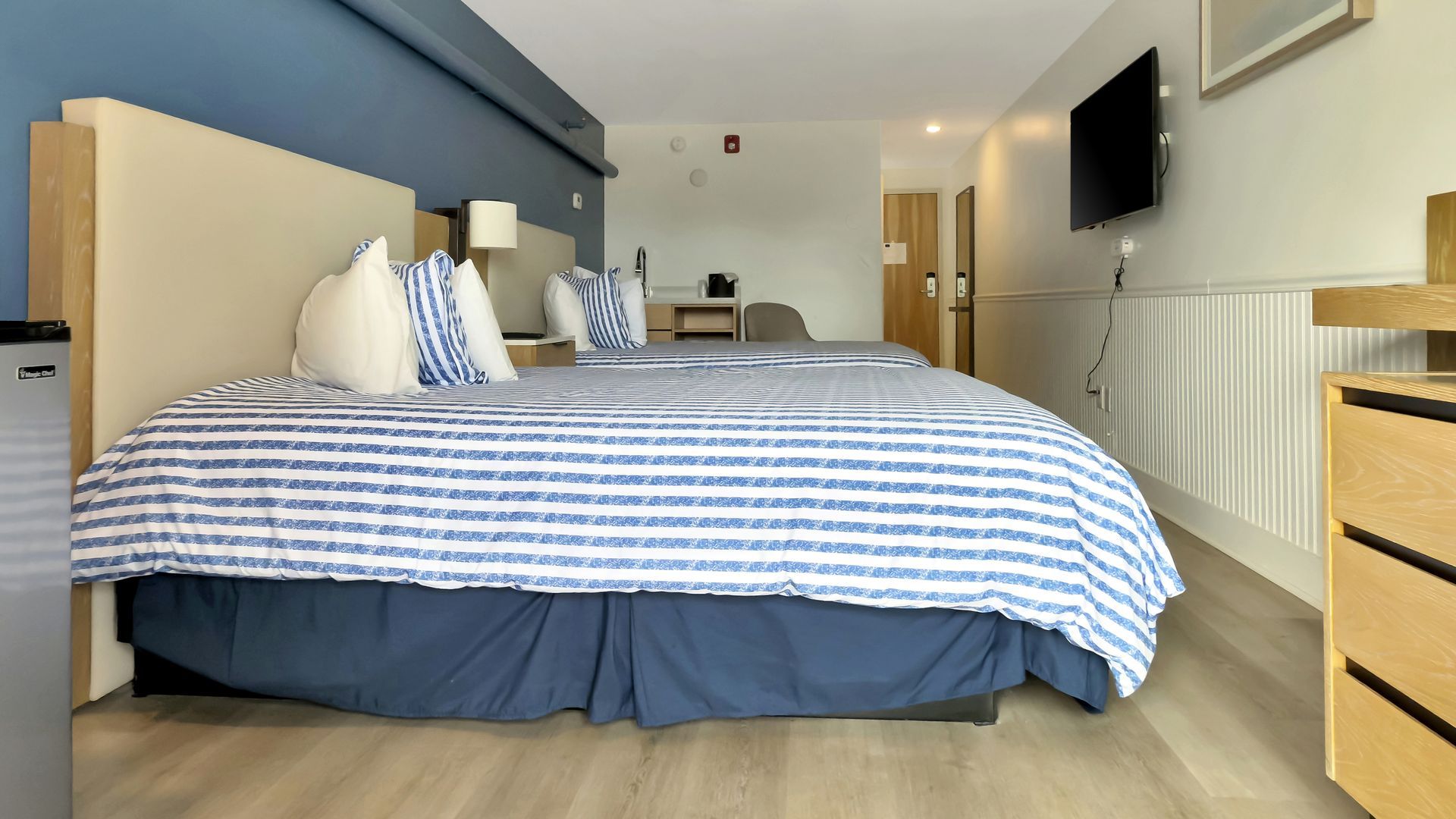 Hotel room with two beds, blue striped comforters, and a wall-mounted TV.