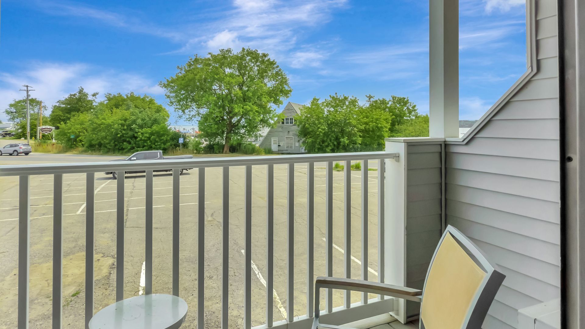 Balcony overlooking a parking lot, trees, and sky. A chair and table sit on the balcony.