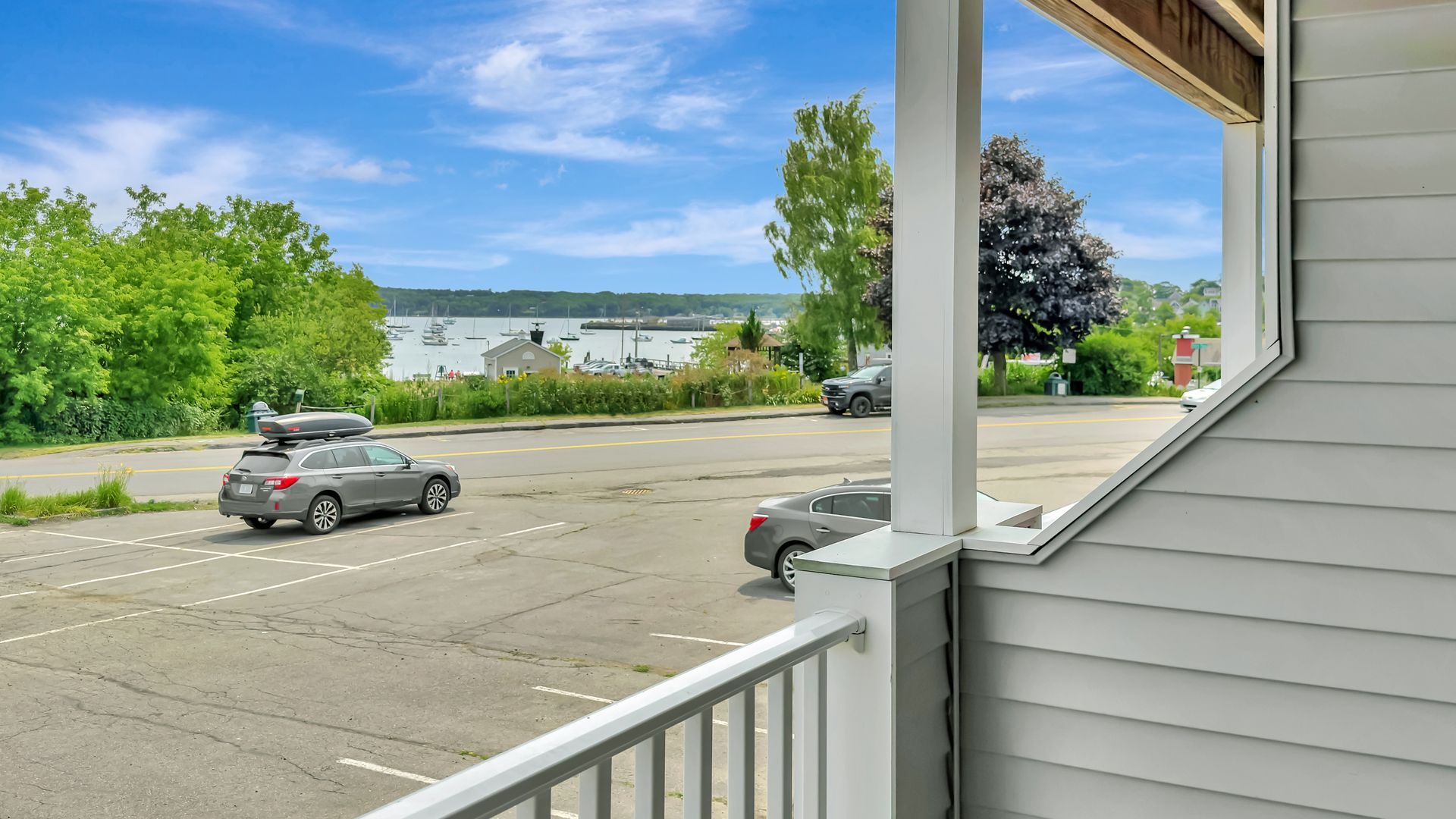 View from a porch overlooking a parking lot and water, with cars, trees, and boats.