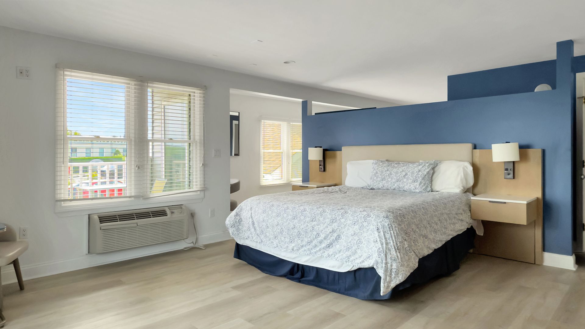 Bright hotel room with king bed, blue headboard, and windows overlooking a city street.