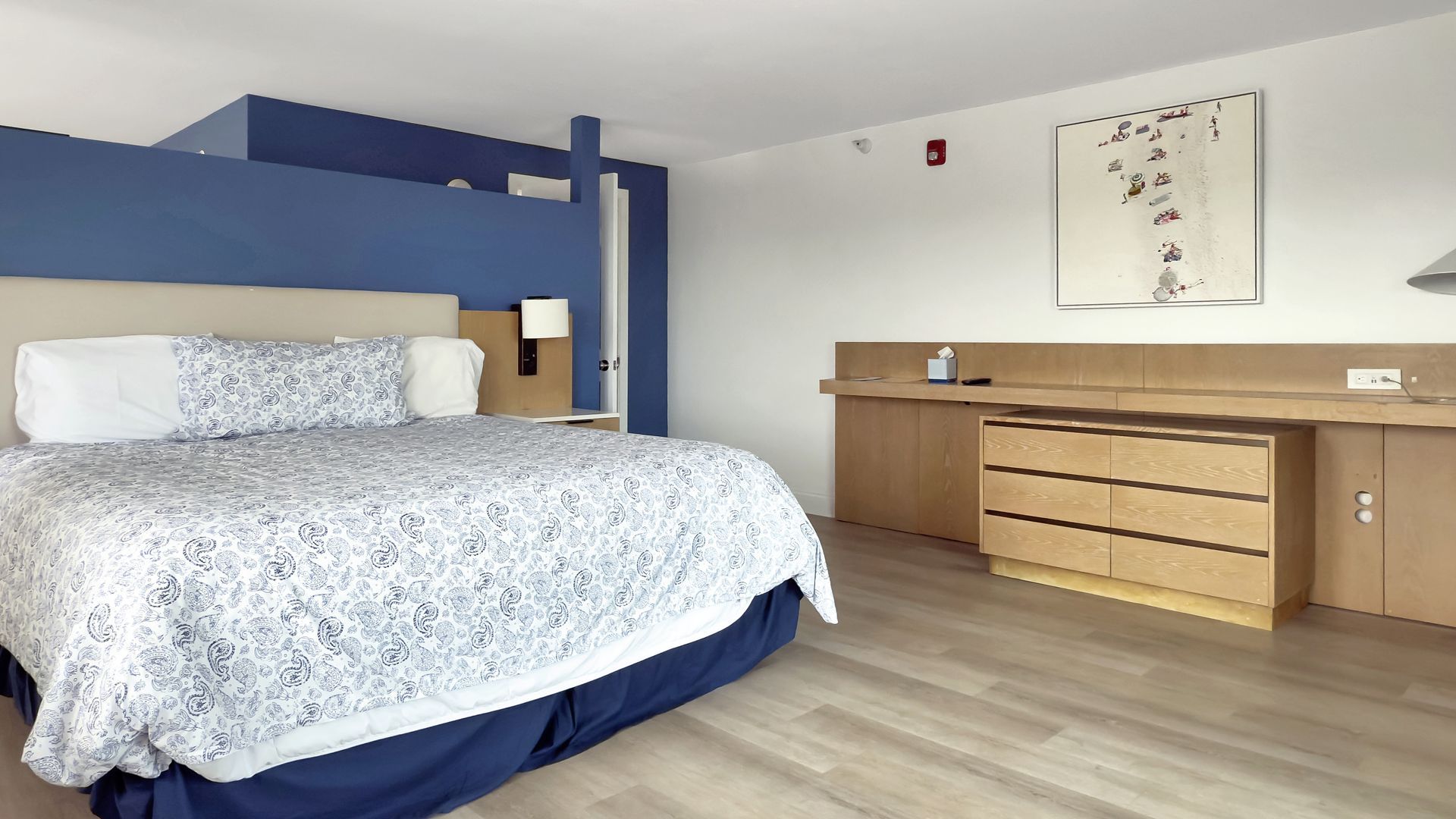 Hotel bedroom with a king-size bed, blue headboard, light wood furniture, and a painting on the wall.