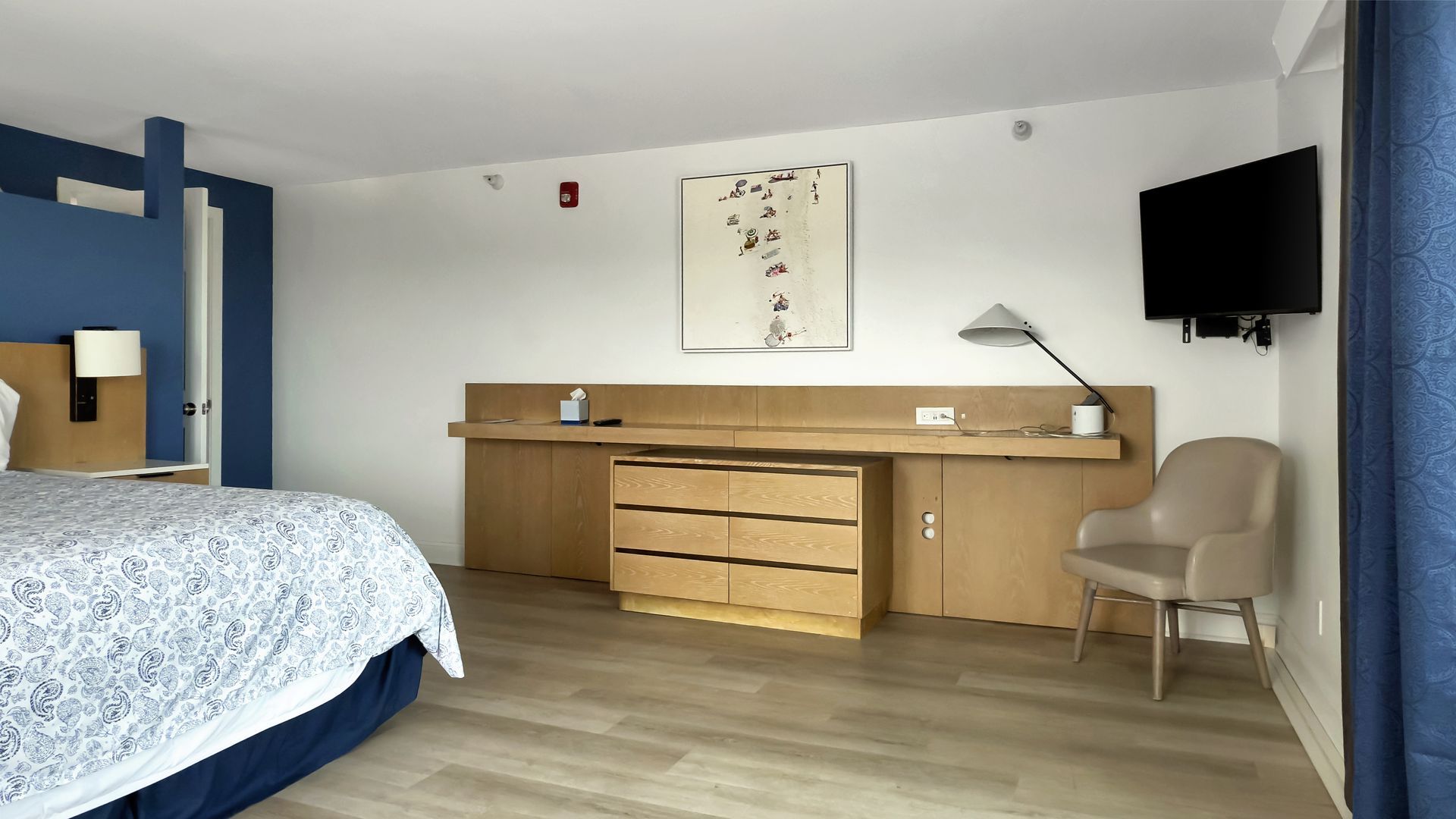 Hotel room with a bed, dresser, TV, and chair. Light wood furniture against white walls.