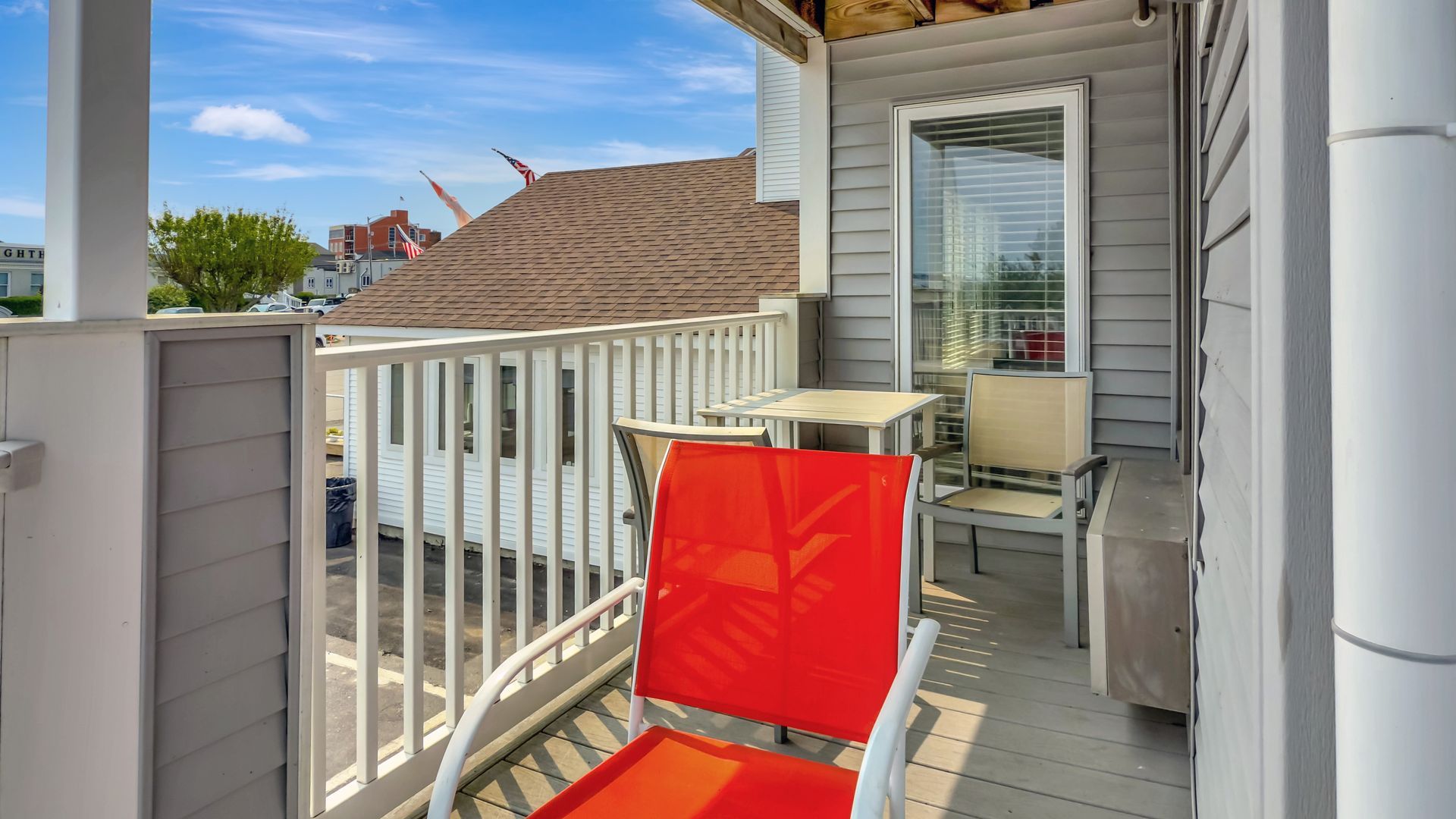 Balcony with red chair and small table, overlooking neighborhood with blue sky.