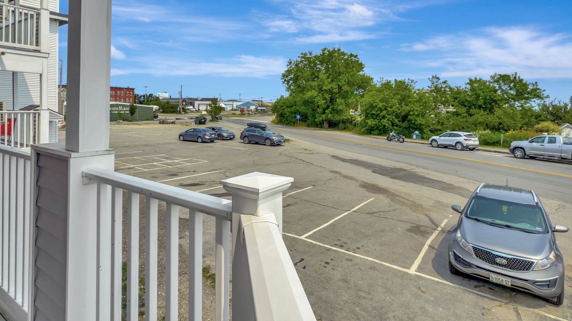 View from a porch of a parking lot with cars, buildings, trees, and blue sky.