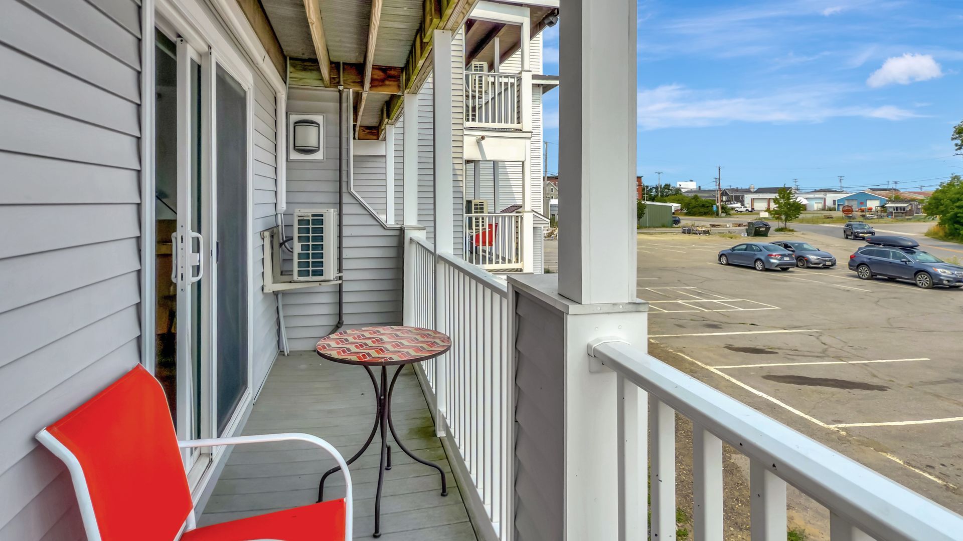 Balcony with red chair, small table, overlooking a parking lot and buildings on a sunny day.
