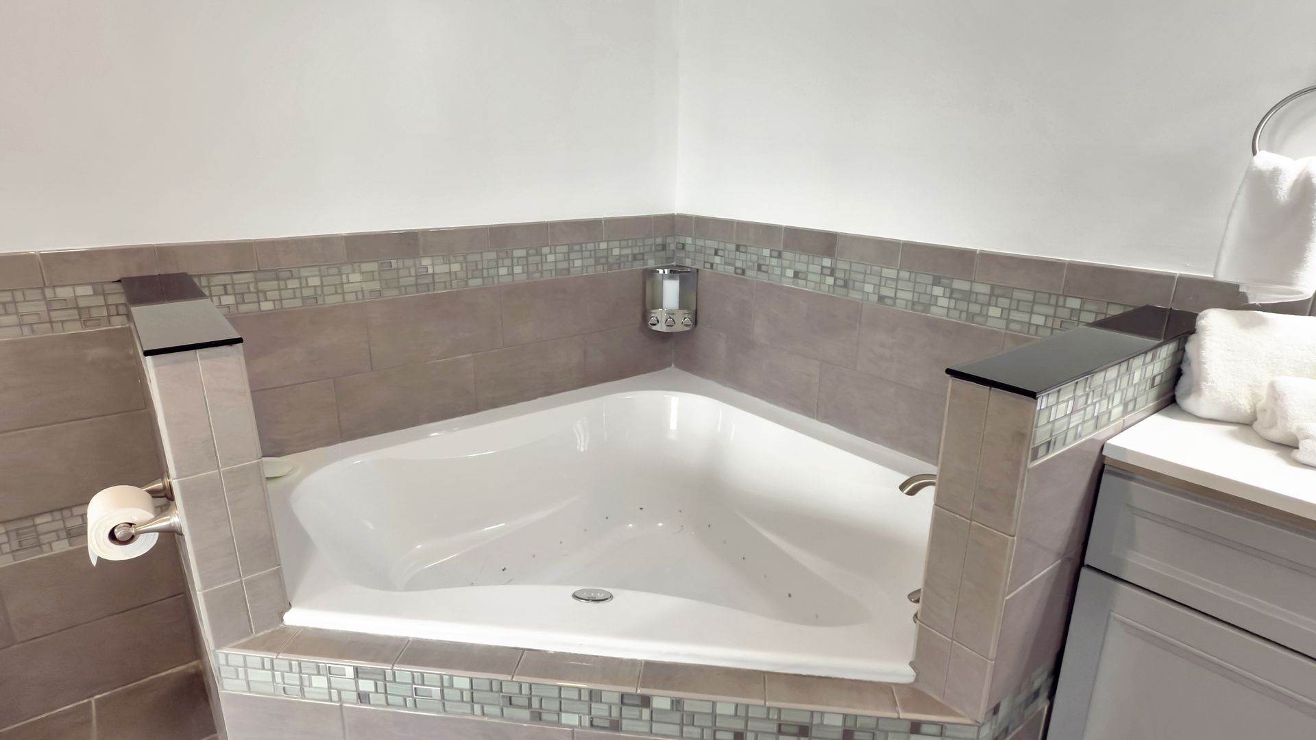 Corner jacuzzi tub surrounded by gray tile and mosaic trim in a bathroom.