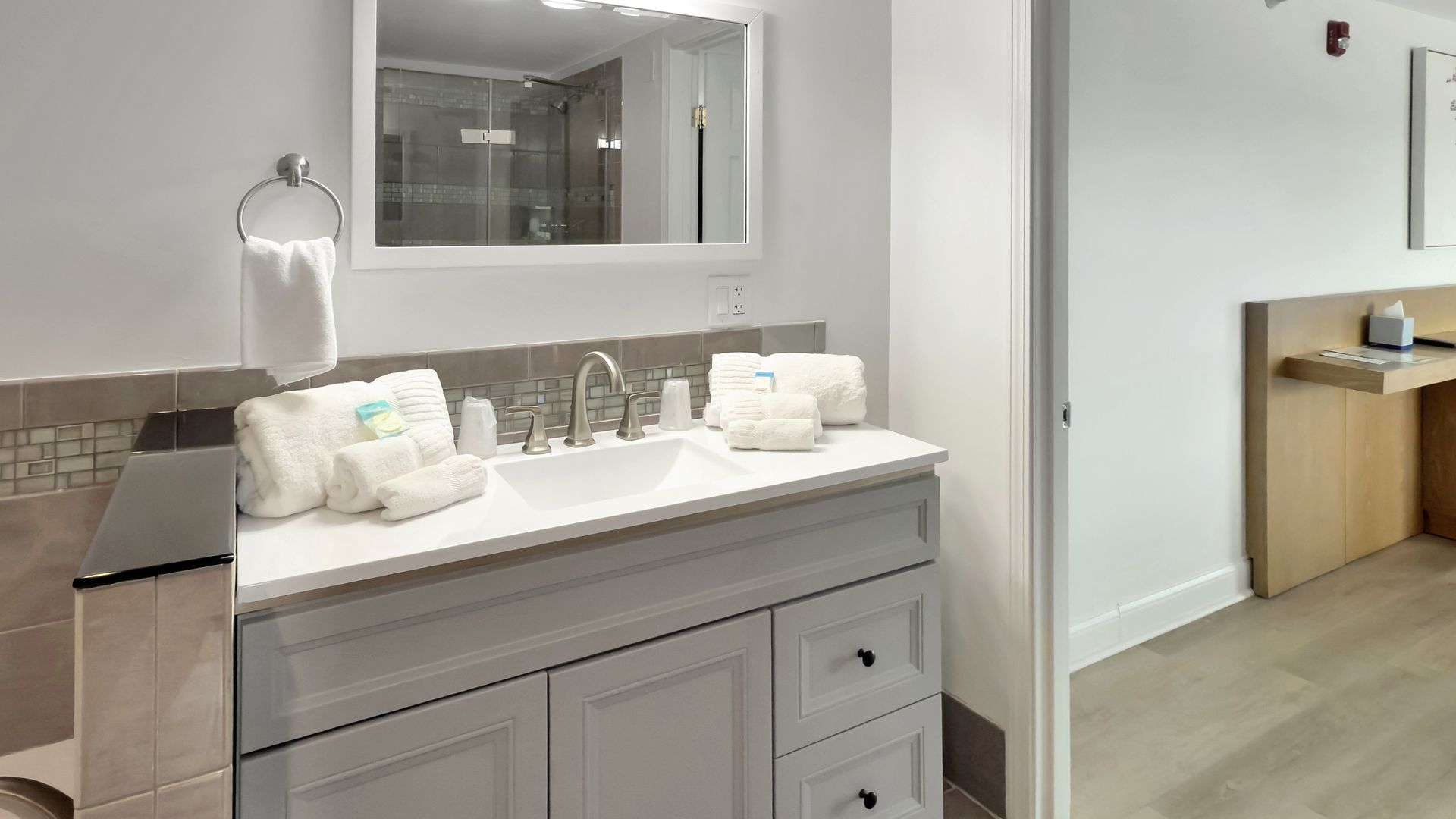 Bathroom with gray vanity, white sink, mirror, and towels, and a doorway to a neutral-toned room with a desk.