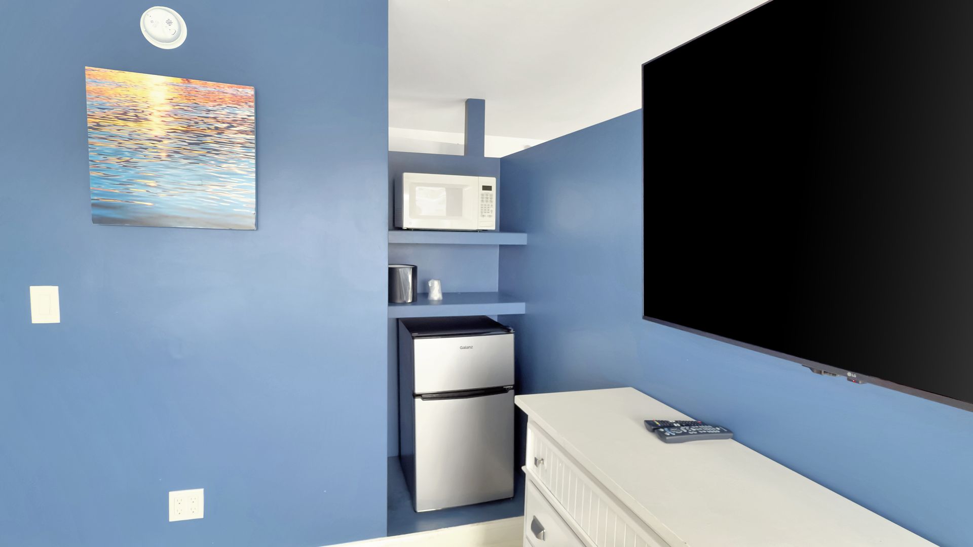 Small kitchenette with microwave, fridge, and TV in a blue-painted room.