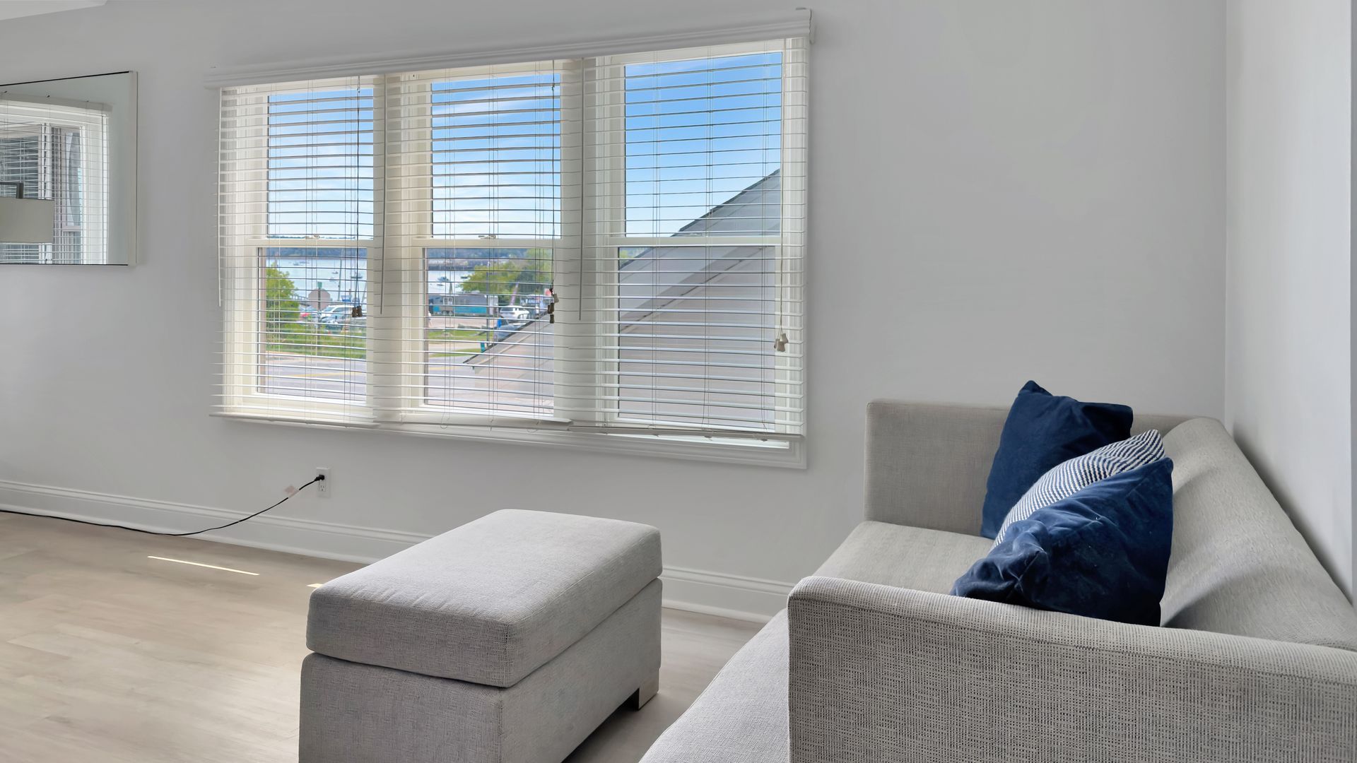 Living room with light gray sofa, ottoman, window with blinds, blue pillows, and light.