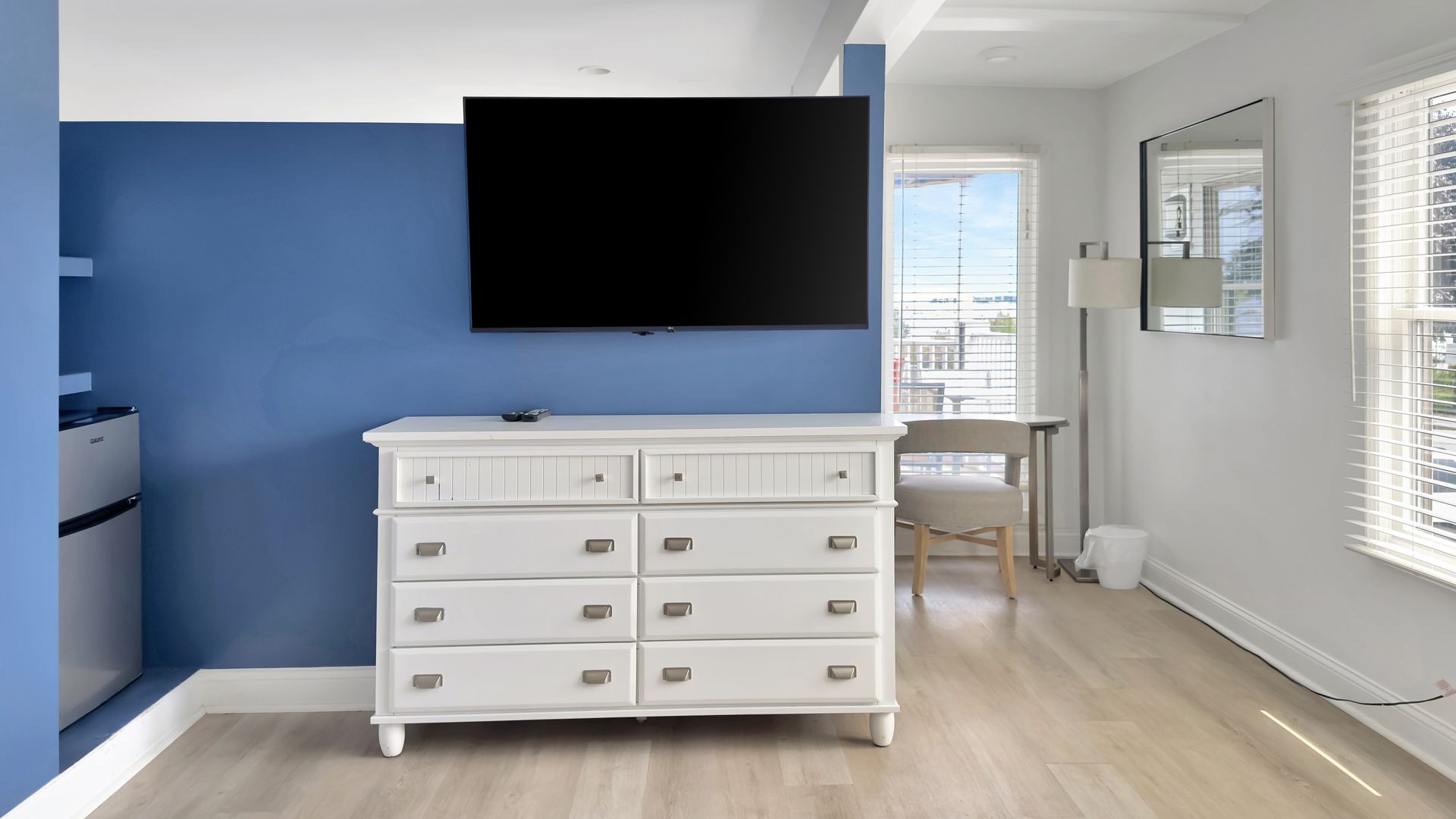 White dresser beneath a large TV on a blue wall. Bright room with a window and desk.