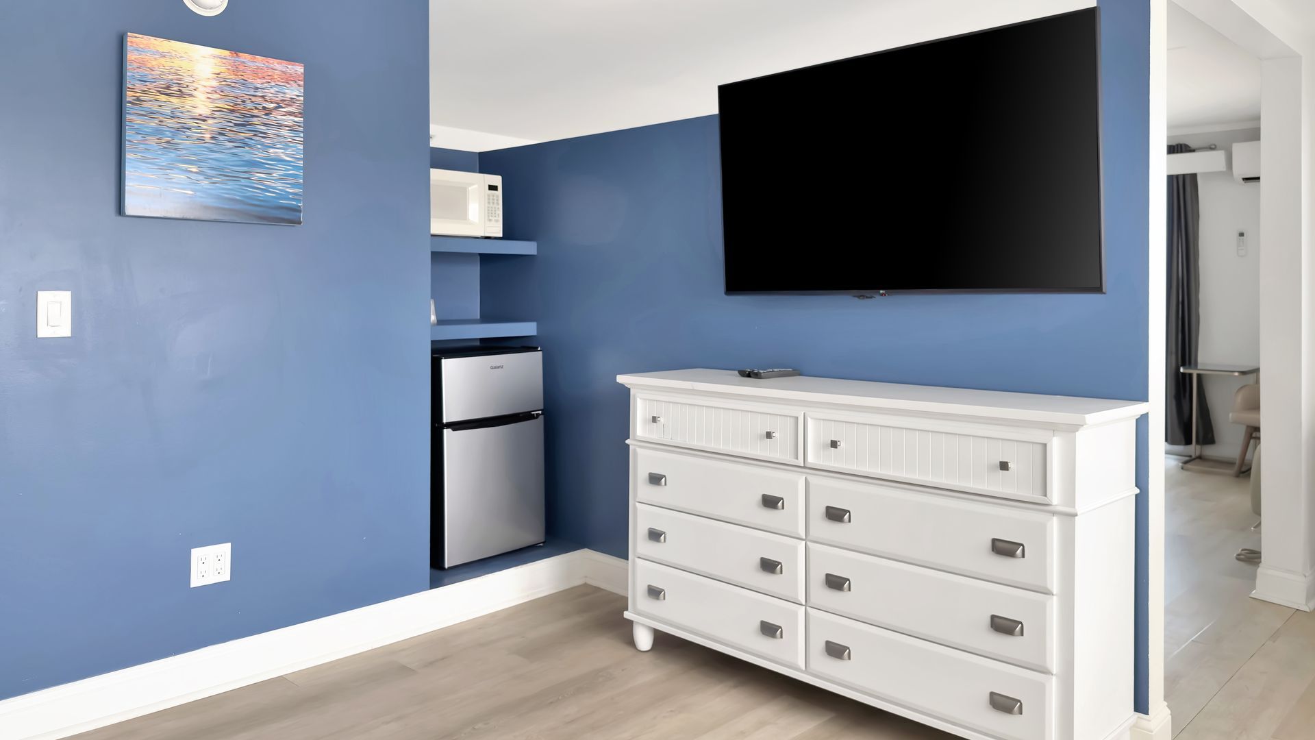 Blue wall with TV, white dresser, small refrigerator, and microwave.