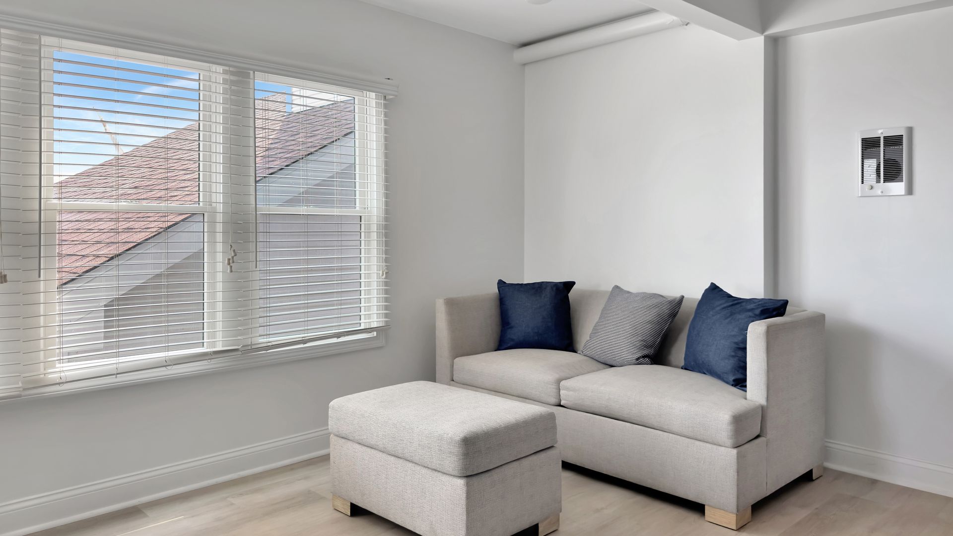 A small, bright living room with a couch and ottoman, beside a window with a view.