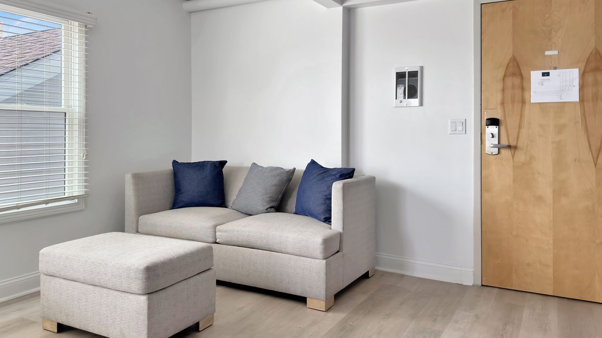 A small, light-colored couch with blue pillows and a matching ottoman, next to a closed wooden door in a room.