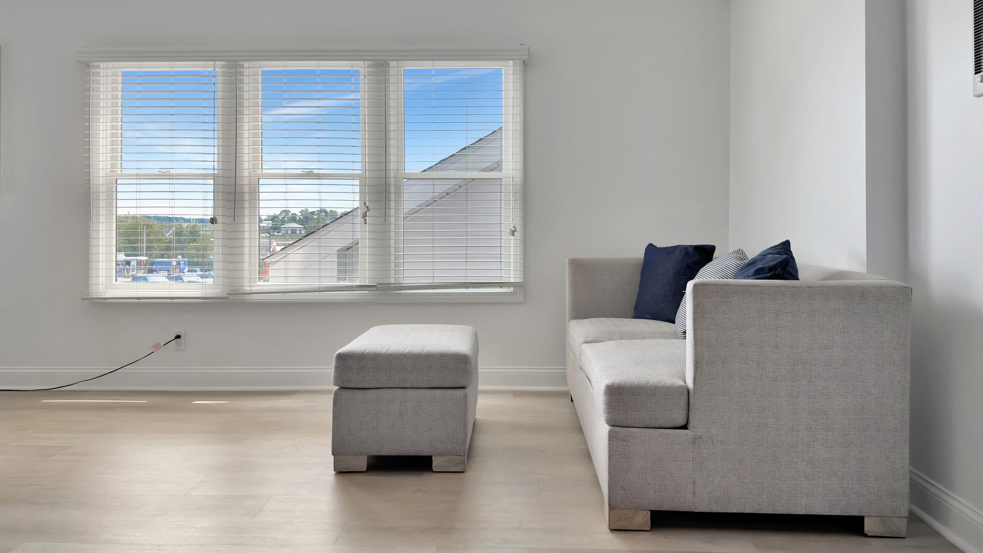 Living room with gray couch, ottoman, and three-panel window with blinds.