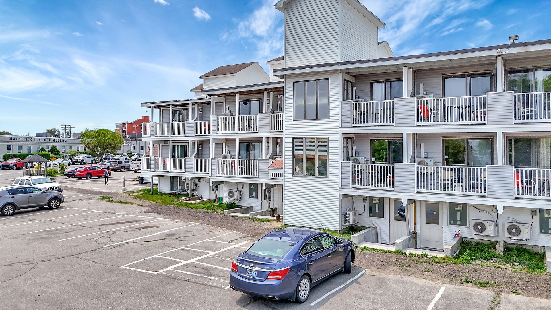 A multi-story white building with balconies and a parking lot with cars on a sunny day.