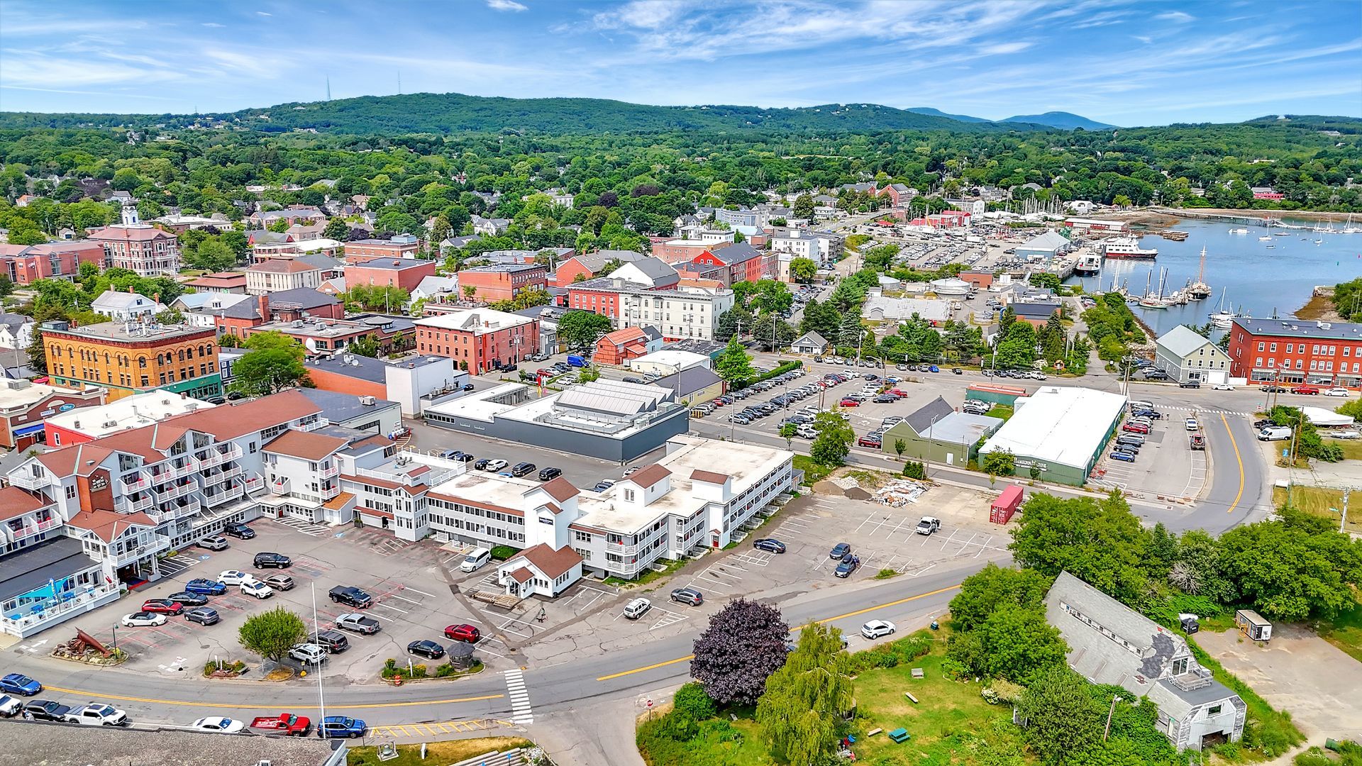 Aerial view of a coastal town with buildings, roads, parking, and a harbor under a blue sky.